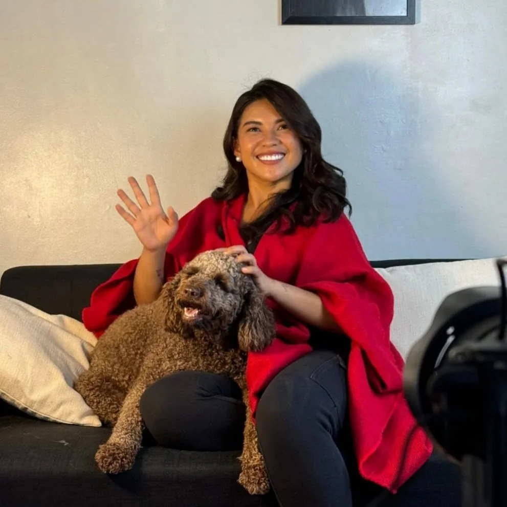 A woman with dark hair, smiling and waving, sitting on a black couch with a brown dog in her lap.