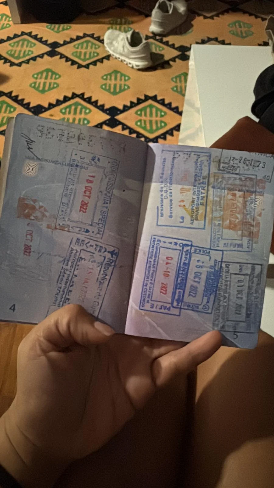An open passport with multiple stamps held by a hand. In the background, there are white sneakers and a patterned mat with green and black geometric designs.