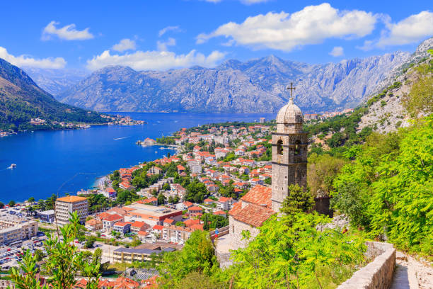 View of a coastal town with red-roofed buildings, a church steeple with a cross, and a large lake surrounded by mountains under a partly cloudy sky.