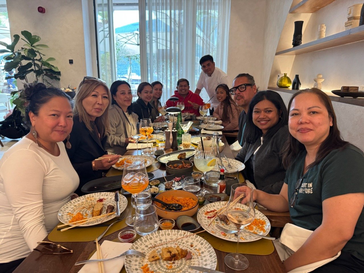 A diverse group of people seated around a long dining table with various partially eaten dishes, drinks, and utensils in a well-lit restaurant with large windows and decorative shelves.