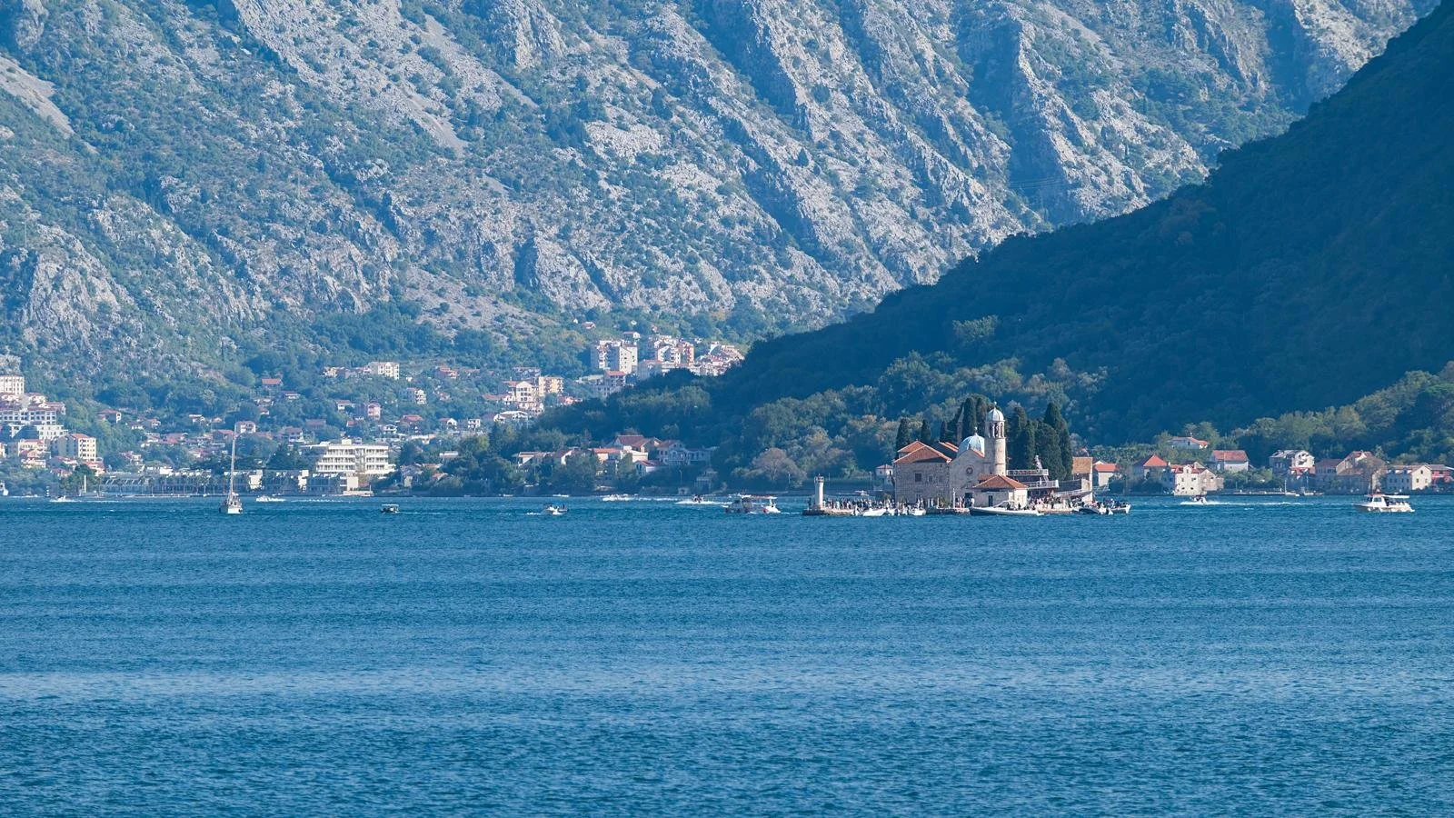Scenic view of a small church on an island surrounded by water called Our Lady of the Rocks in Perast, Montenegro with boats nearby and a mountainous shoreline in the background.