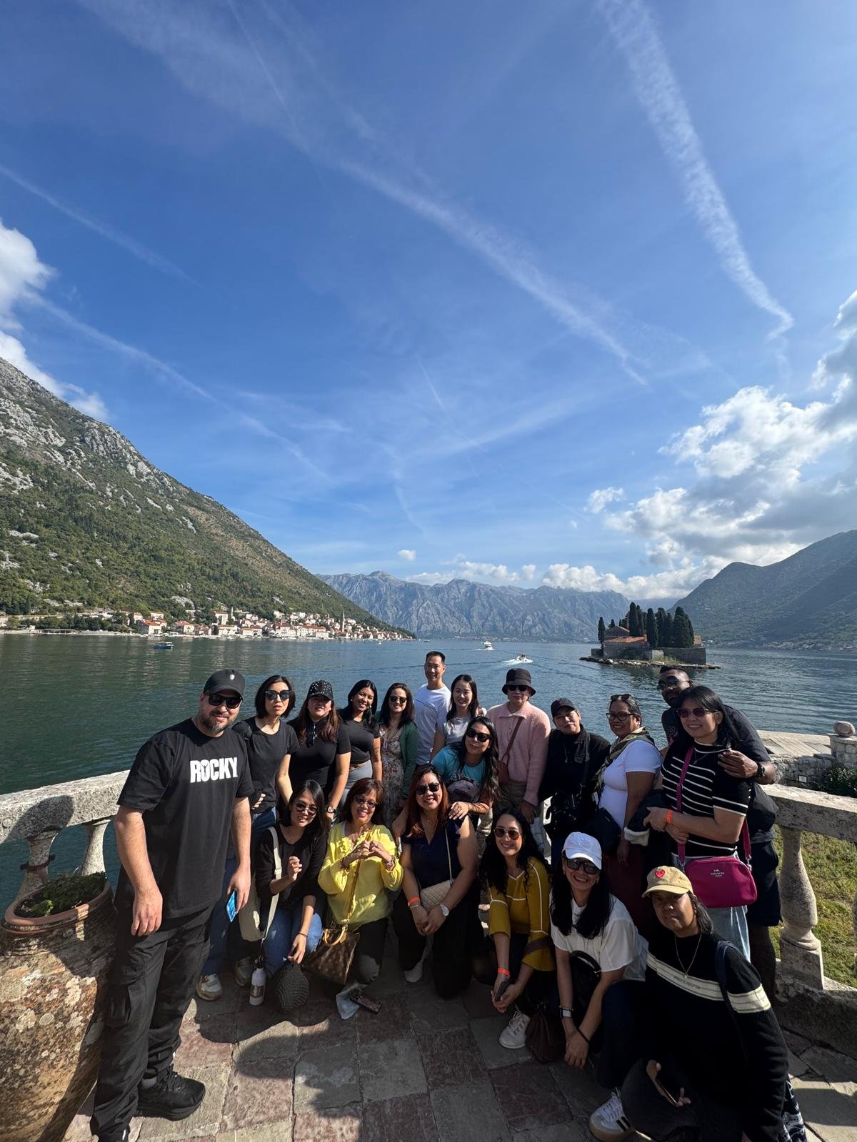 A group of people posing together on a scenic lakeside with mountains in the background, under a blue sky with clouds.