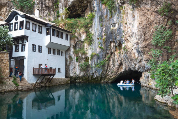A white building with multiple windows situated next to a rocky cliff with a cave opening. The building is beside a calm body of water with a small boat carrying people nearby. Trees and greenery are present around the scene.