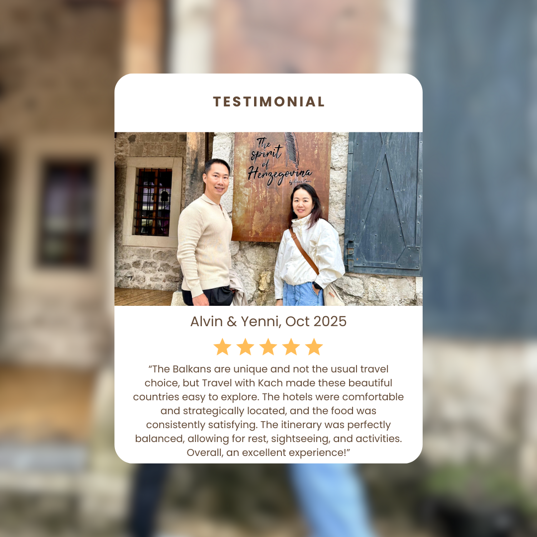 Two people standing in front of a sign that reads 'The Spirit of Herzegovina'. The man is wearing a beige sweater and the woman is wearing a white jacket with a brown crossbody bag.