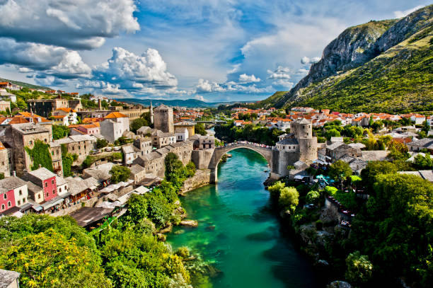 Sunlit view of a historic European town with a stone bridge over a green river, surrounded by colorful buildings and lush greenery, with mountains in the background and a partly cloudy sky.