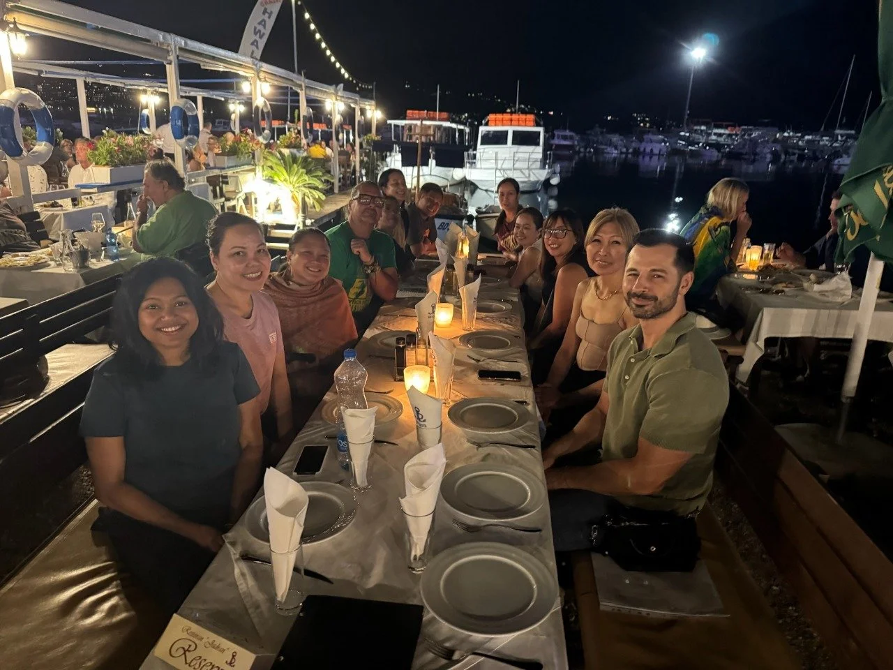 A group of Filipino and American people sitting at a long outdoor dinner table at night, smiling, with a harbor full of boats in the background, under string lights, at a seaside restaurant in Budva, Montenegro