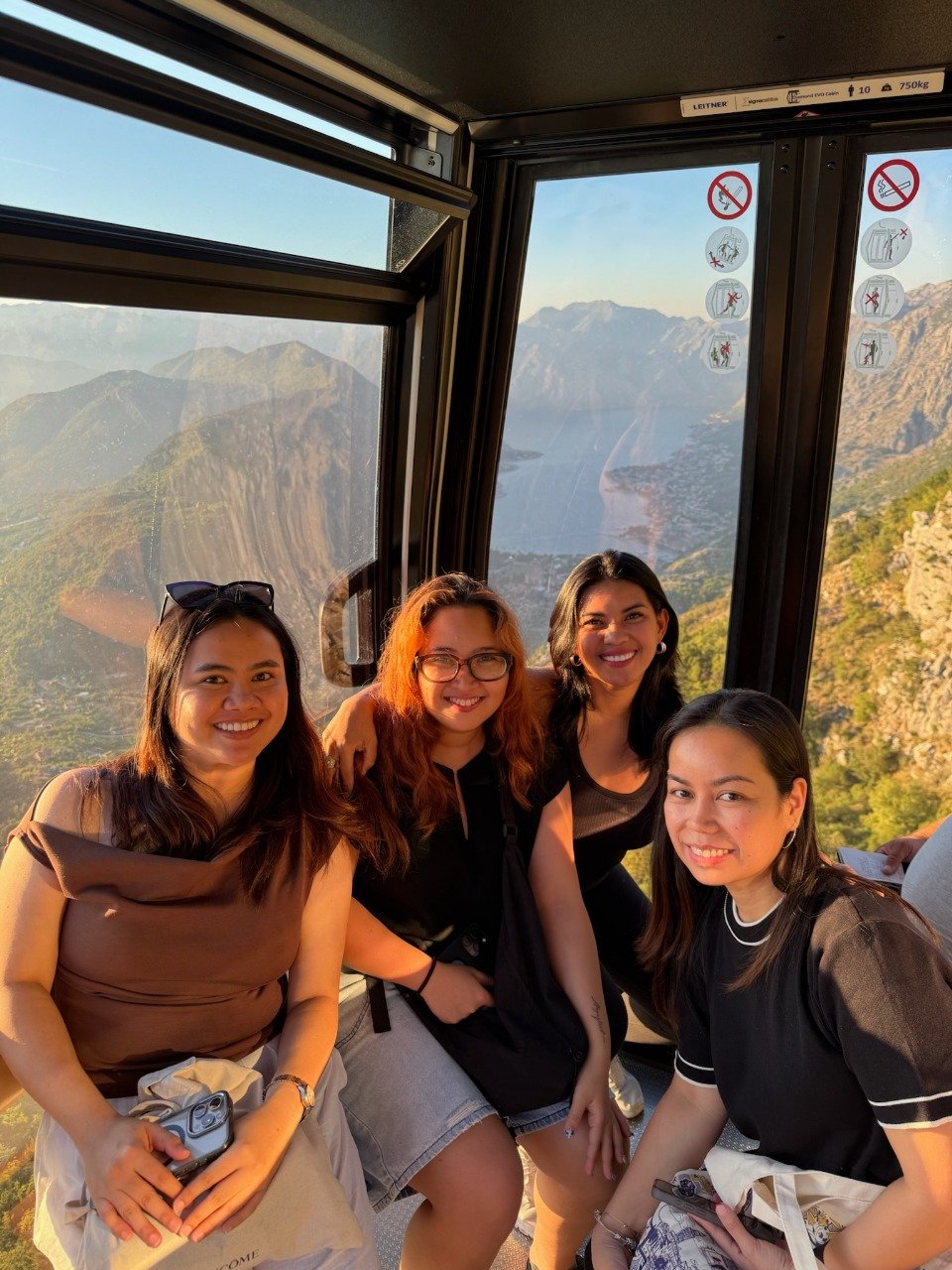 Four women smiling inside a cable car with a mountain and lake view in the background in Kotor, Montenegro.