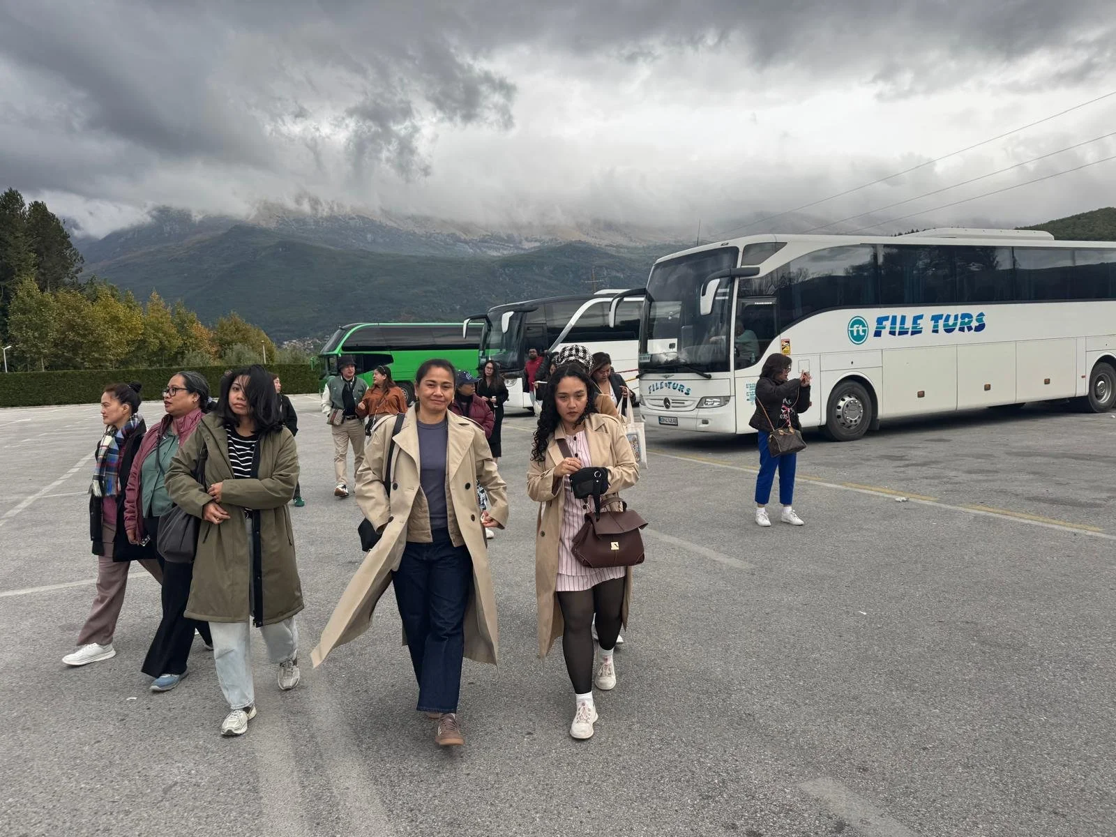 Group of people walking in a parking lot with mountains in the background, several buses parked nearby, and cloudy sky overhead.