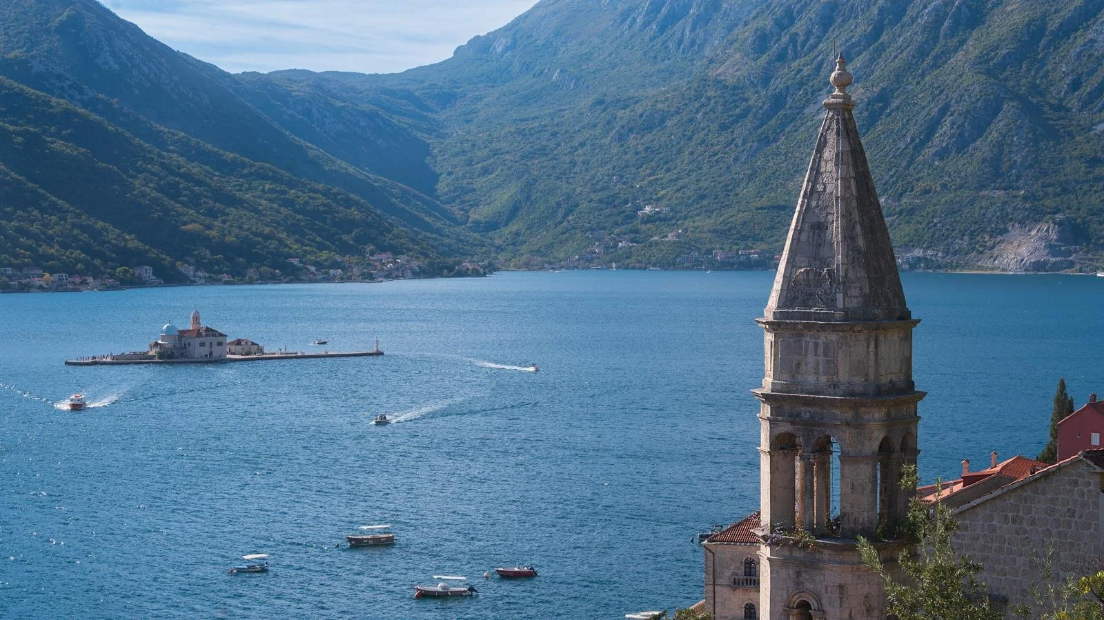 View of a serene seascape with a small fortress on an island and a tall church steeple in the foreground, surrounded by lush green mountains in Perast, Montenegro.