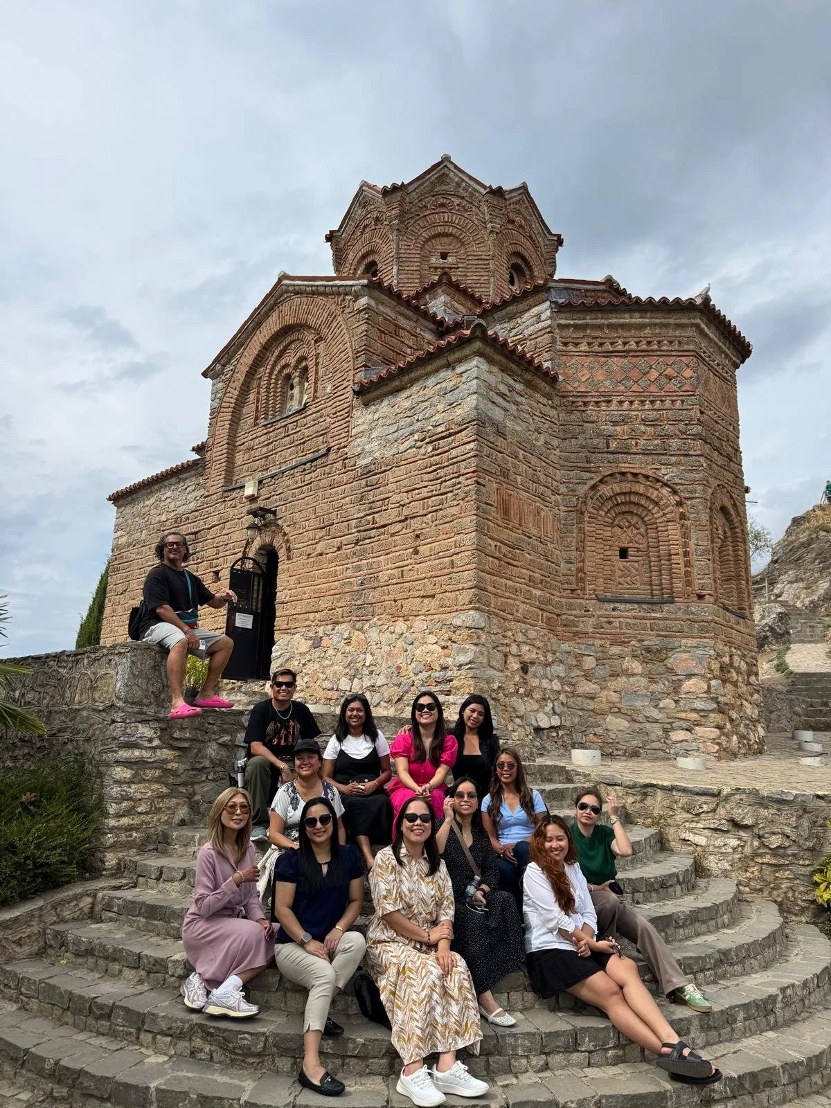 Group of people sitting on stone steps in front of an ancient brick church with a cloudy sky.