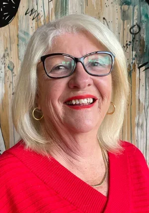 A woman with shoulder-length black hair smiling outdoors, wearing a blue top with a gathered neckline, surrounded by green plants.