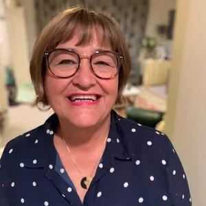 A man with glasses, a nose piercing, and a beard, wearing a dark green sweater, sitting indoors with a blurred background.