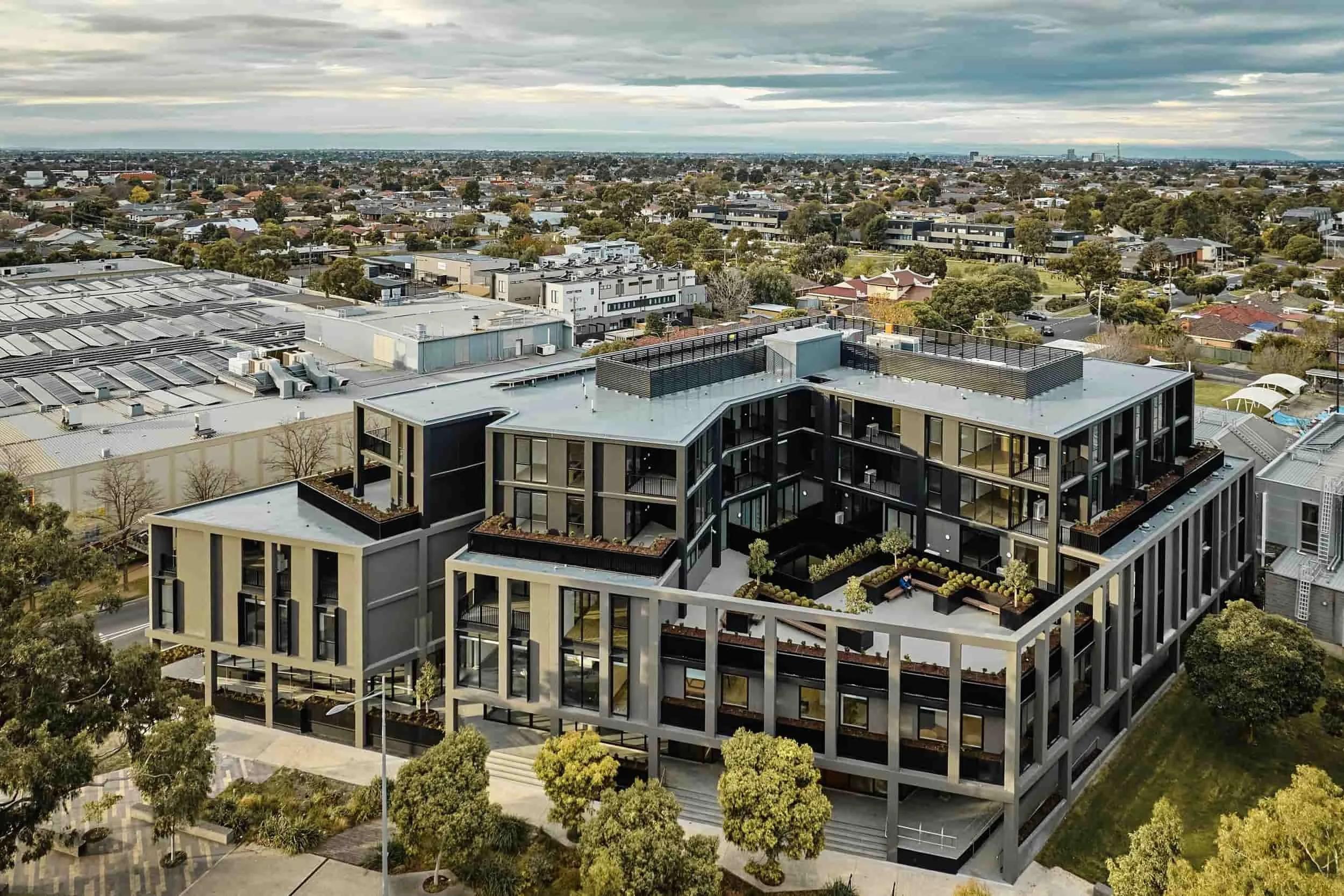 An aerial view of a modern, multi-story residential building with balconies and rooftop gardens in a suburban area with other buildings and trees in the background.