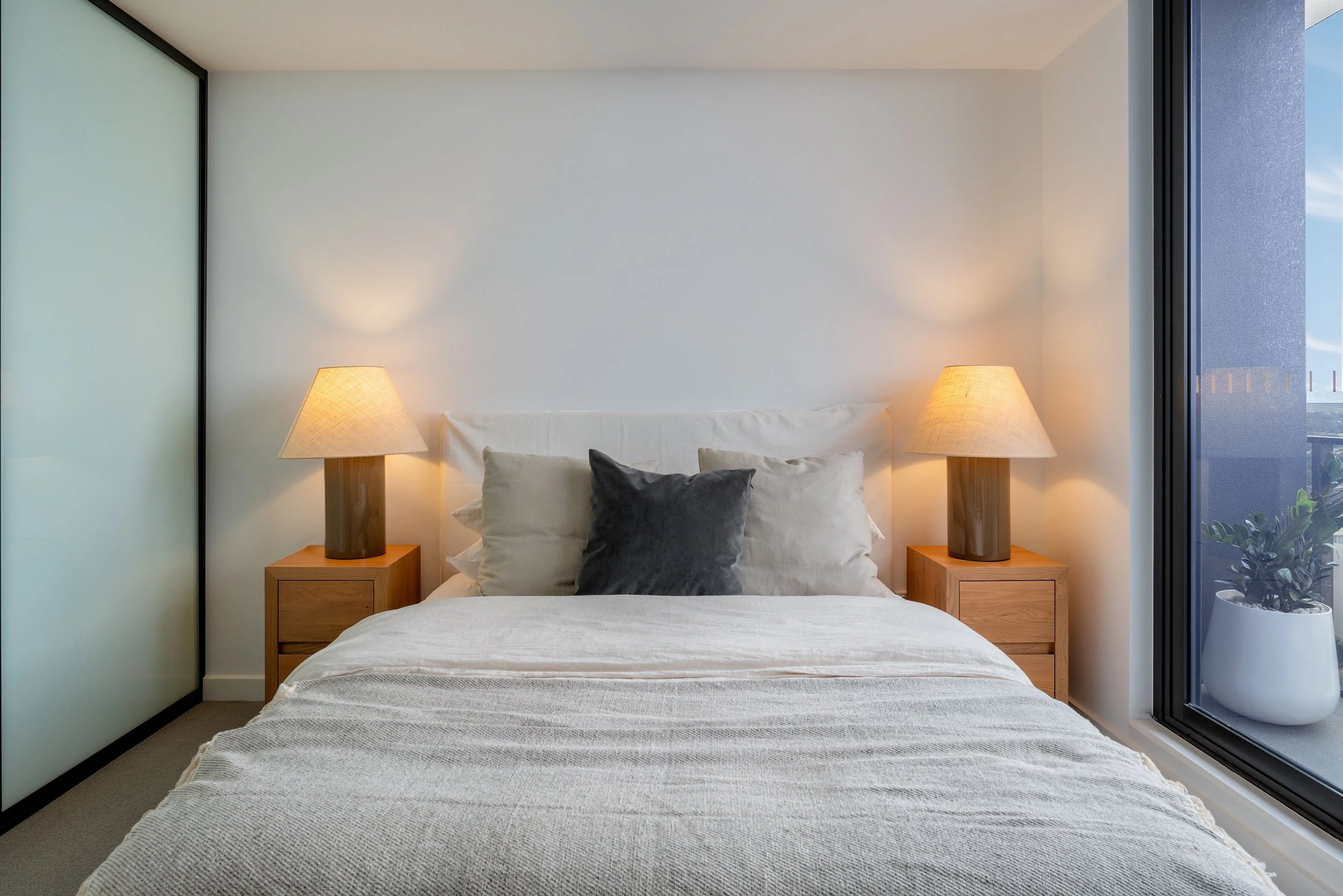 Modern bedroom with a neatly made bed, two bedside tables with lamps, and a large window letting in natural light.