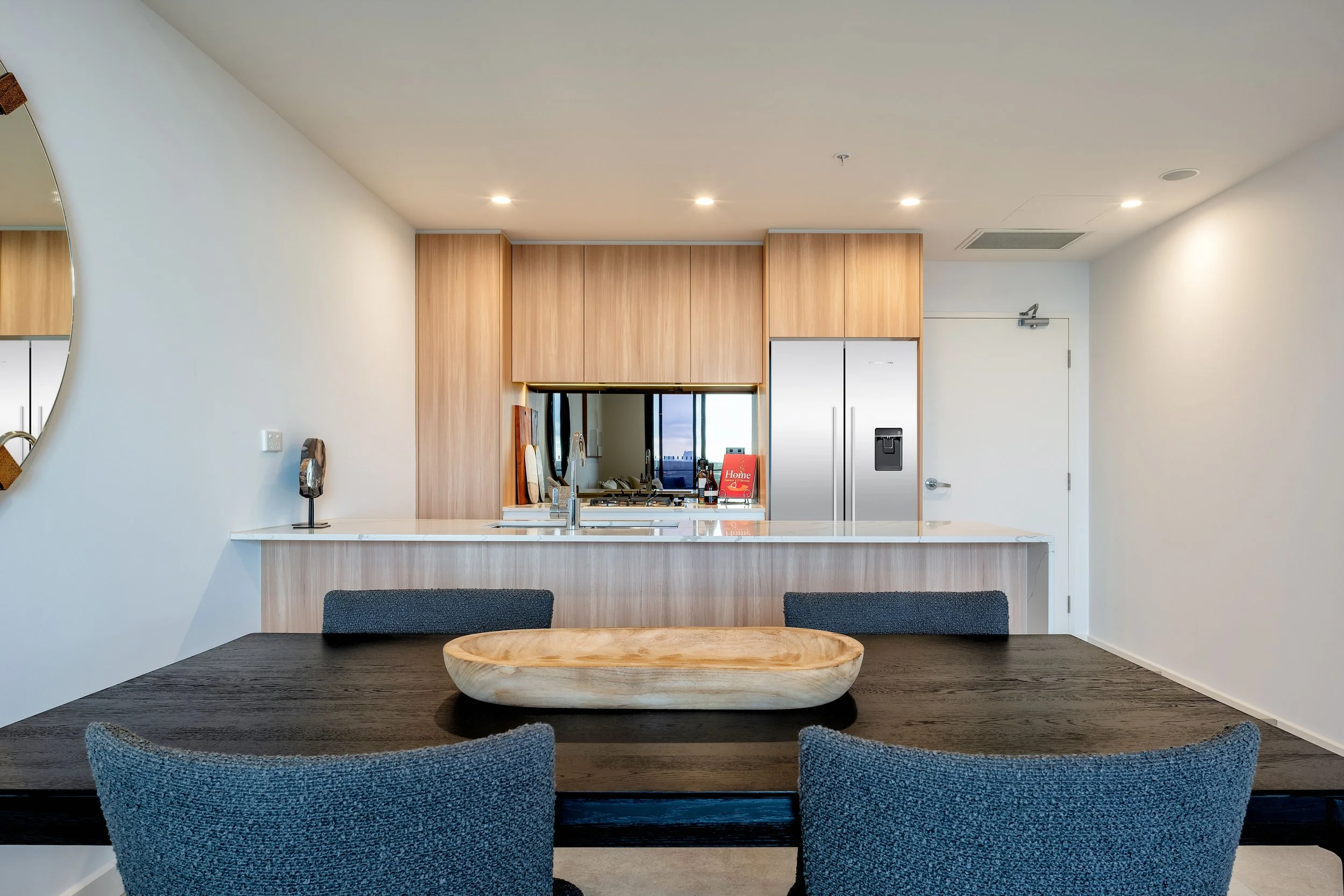 Modern kitchen with wood cabinets, white countertops, stainless steel fridge, and a dark dining table with a wooden bowl centerpiece.