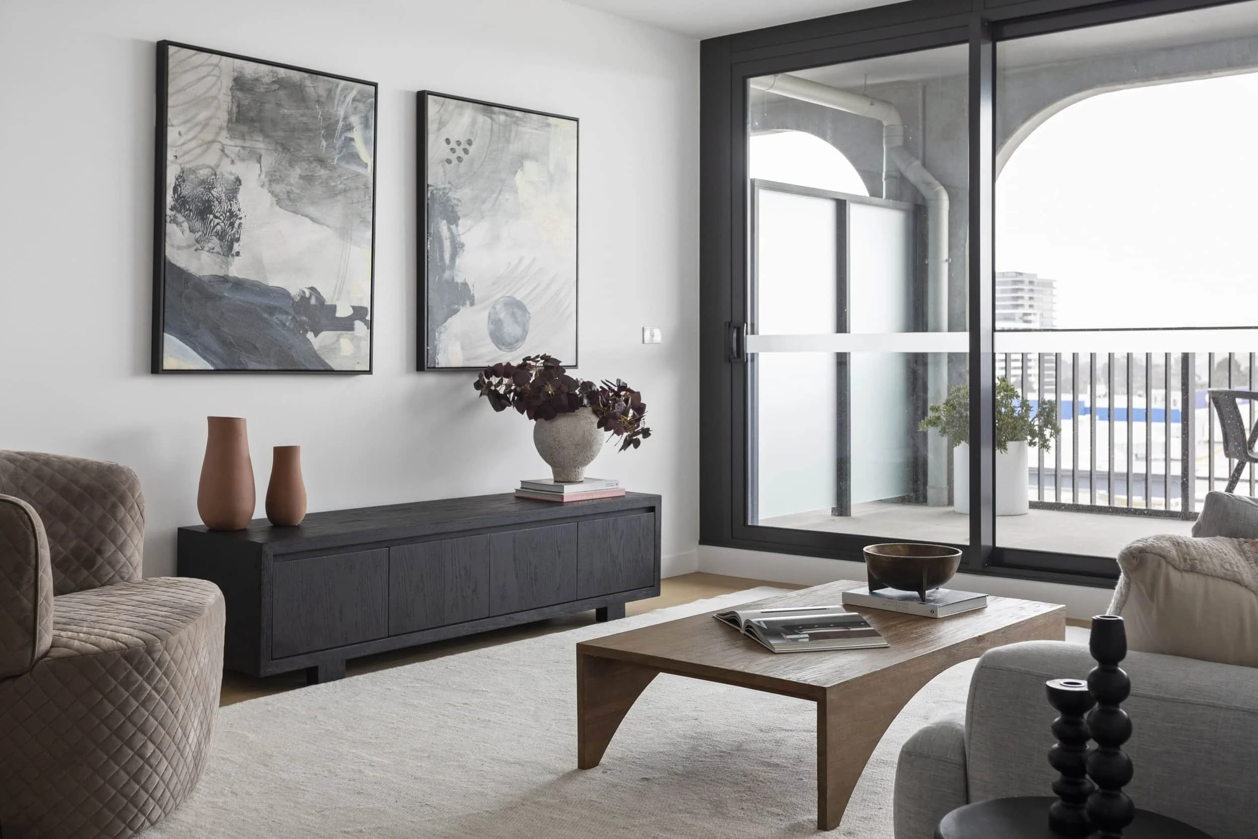 Modern living room with abstract black and white artwork on the wall, a beige quilted armchair, a black sideboard with vases and a flower arrangement, a wooden coffee table with magazines and a bowl, and sliding glass doors leading to a balcony with city view and outdoor furniture.