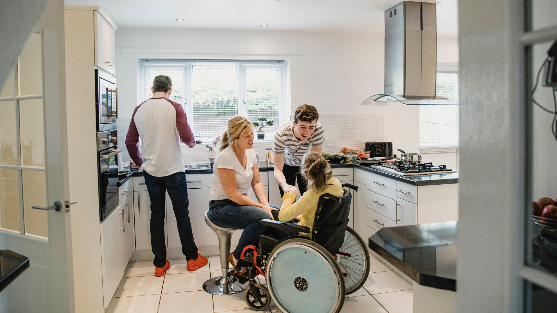 Family in a kitchen with a young girl in a wheelchair, a woman seated talking to her, and a man smiling and holding her hand, with another person cooking in the background.