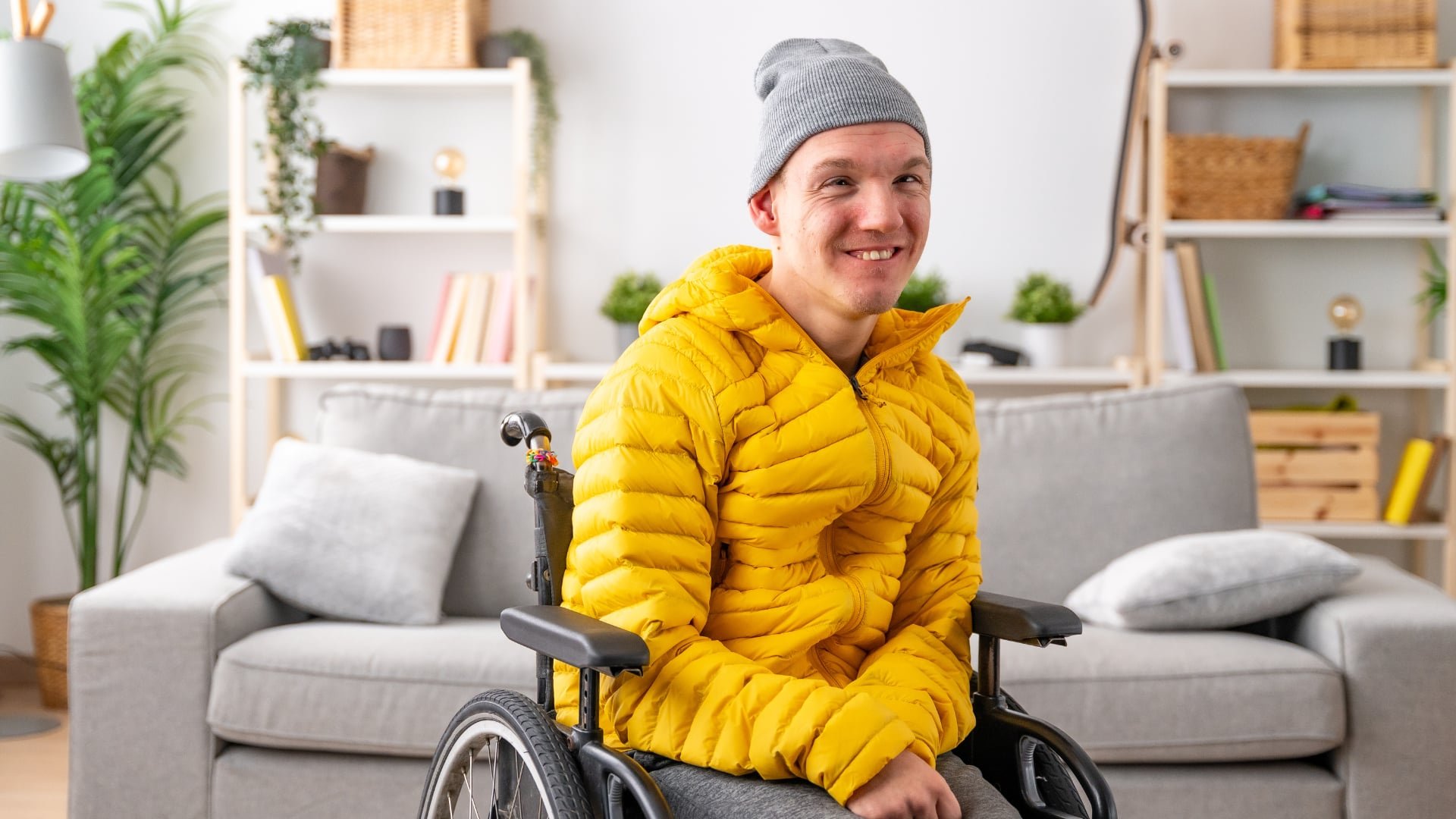 Man in a wheelchair wearing a yellow puffy jacket and a gray beanie, smiling in a cozy, modern living room with a gray couch, pillows, and wooden shelves with plants, books, and decorative items.
