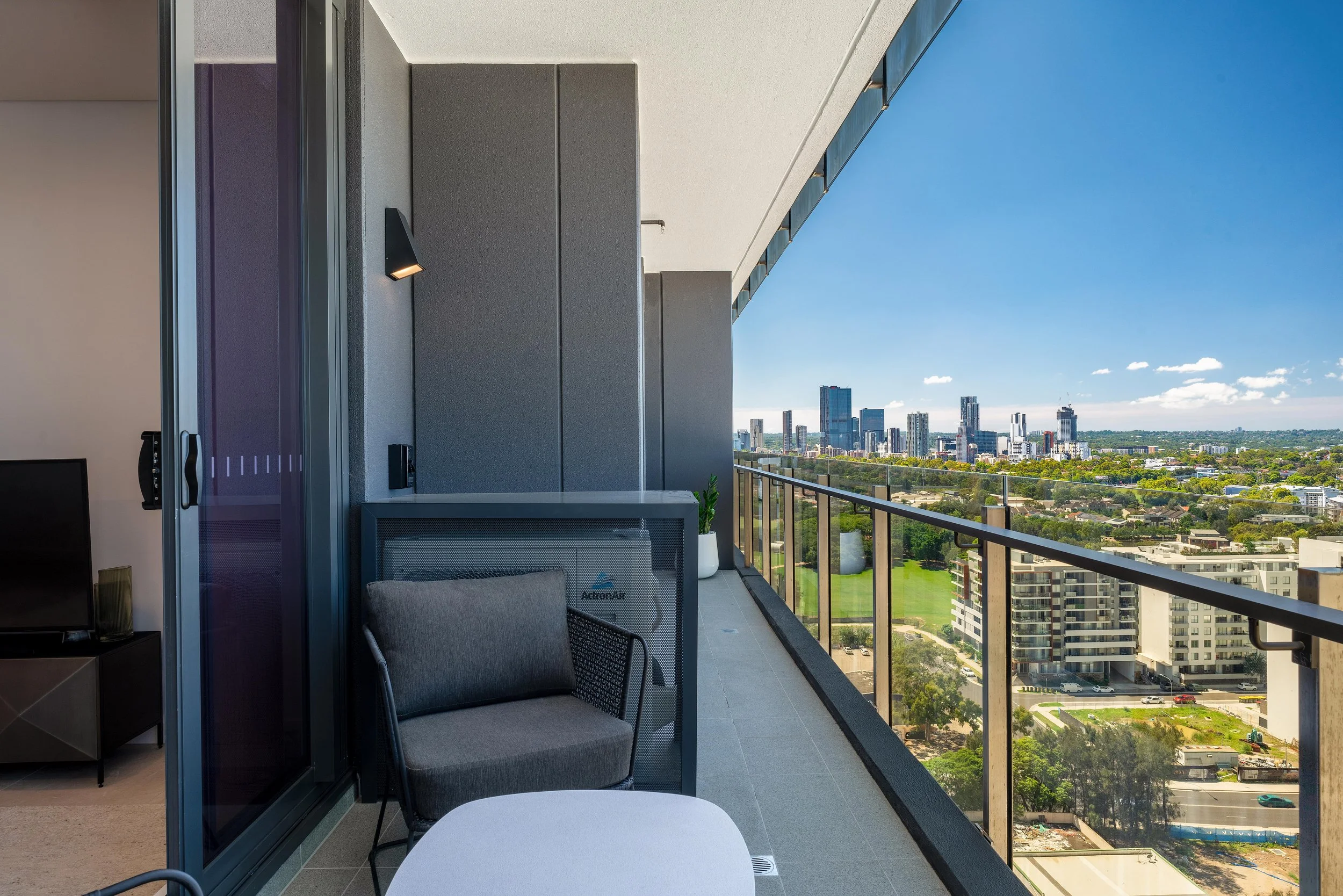 Modern apartment balcony with seating, glass railing, and city skyline views on a sunny day.