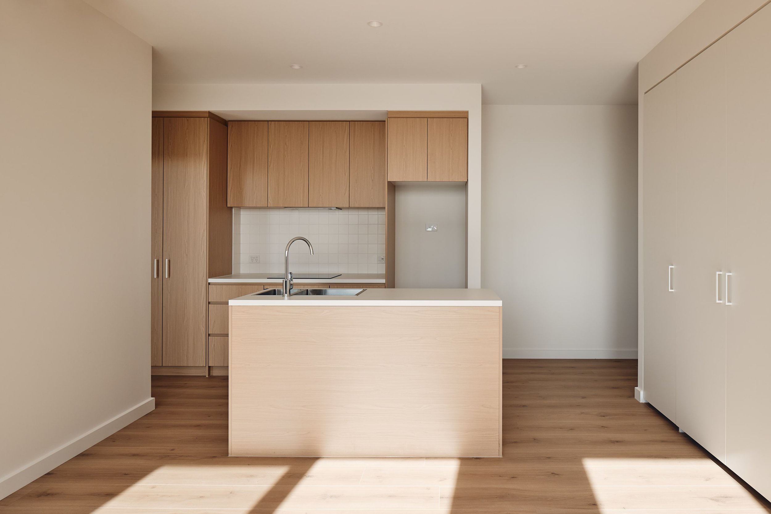 Minimalist kitchen with light wood cabinets, center island with sink, built-in fridge, and white tile backsplash.