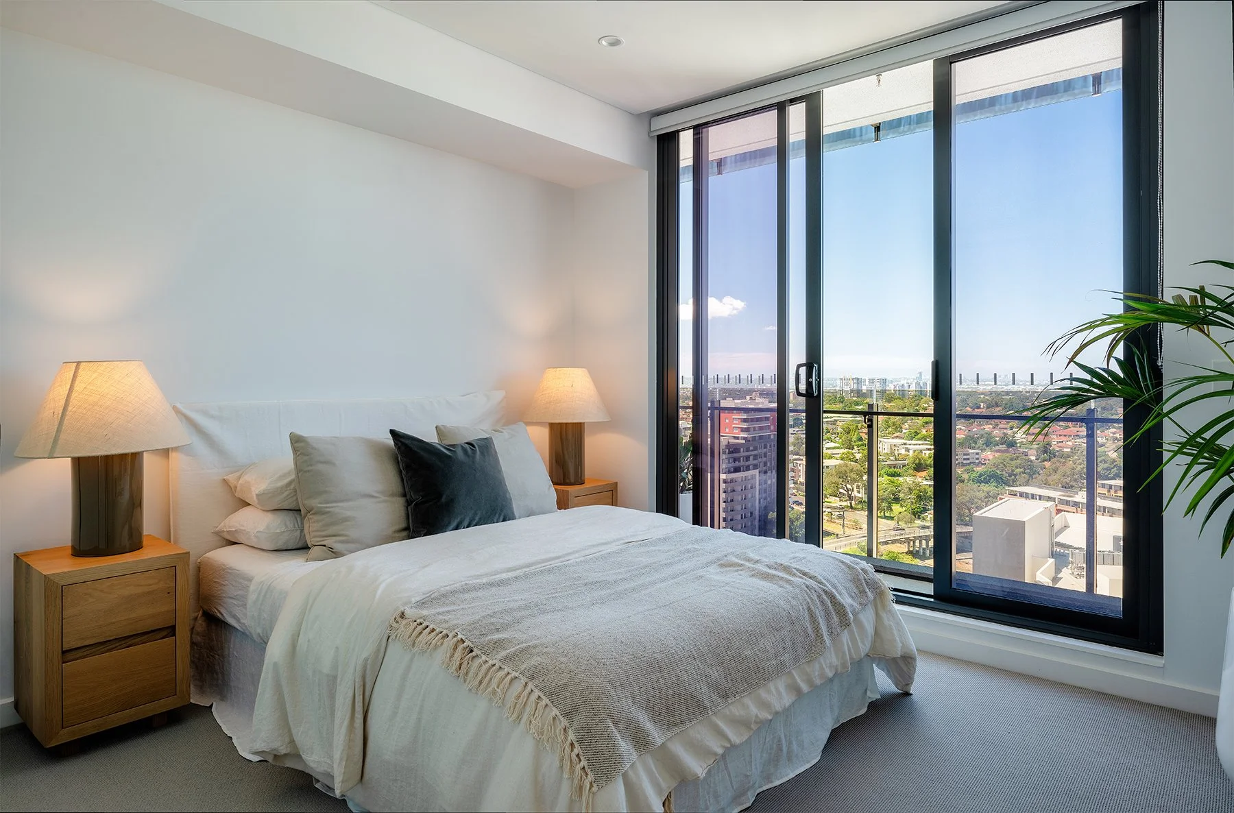 Bright bedroom with a double bed, bedside tables, lamps, and large glass doors opening to a city view balcony.