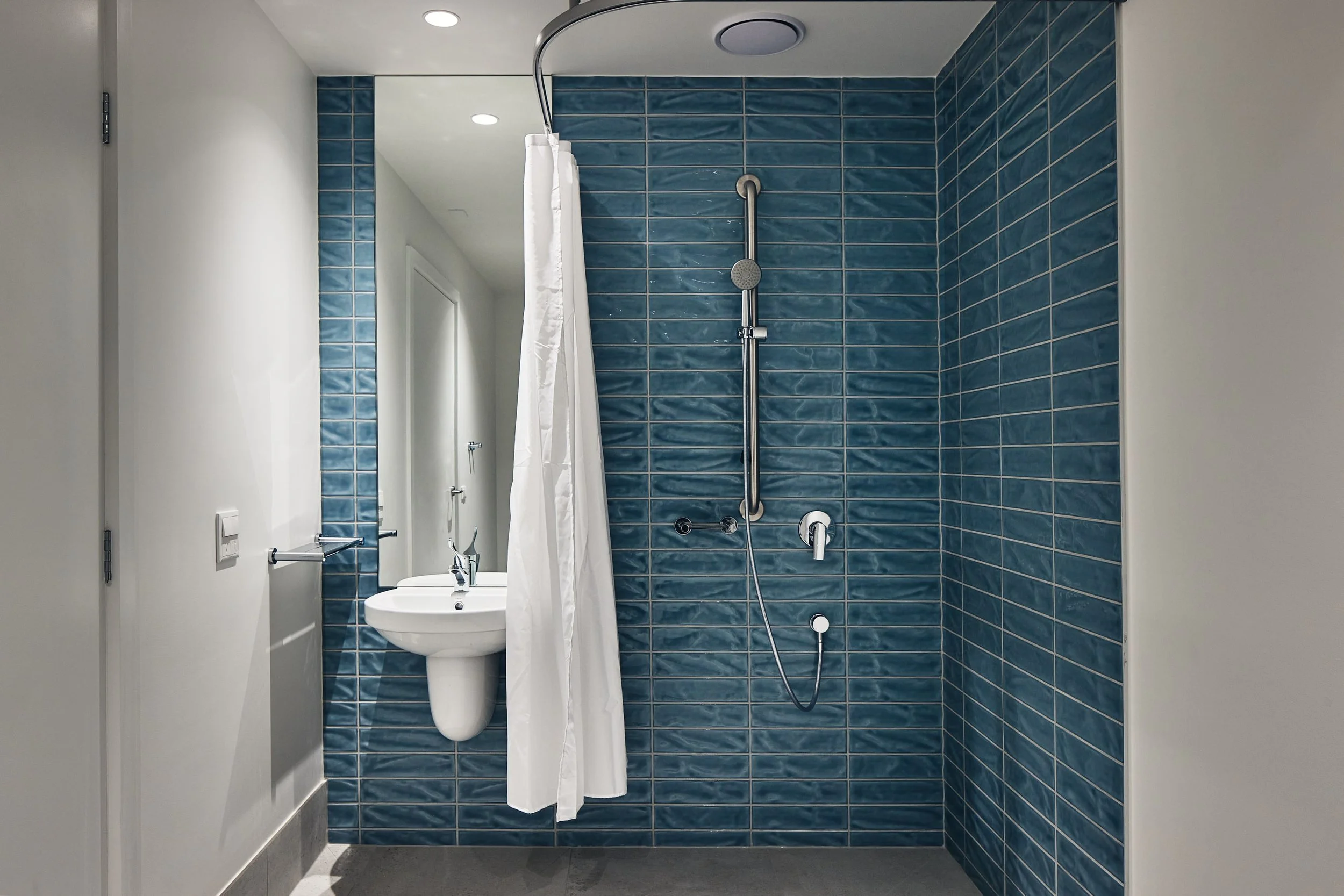Accessible bathroom with blue tiled shower, white curtain, handheld showerhead, grab bar, and wall-mounted sink.