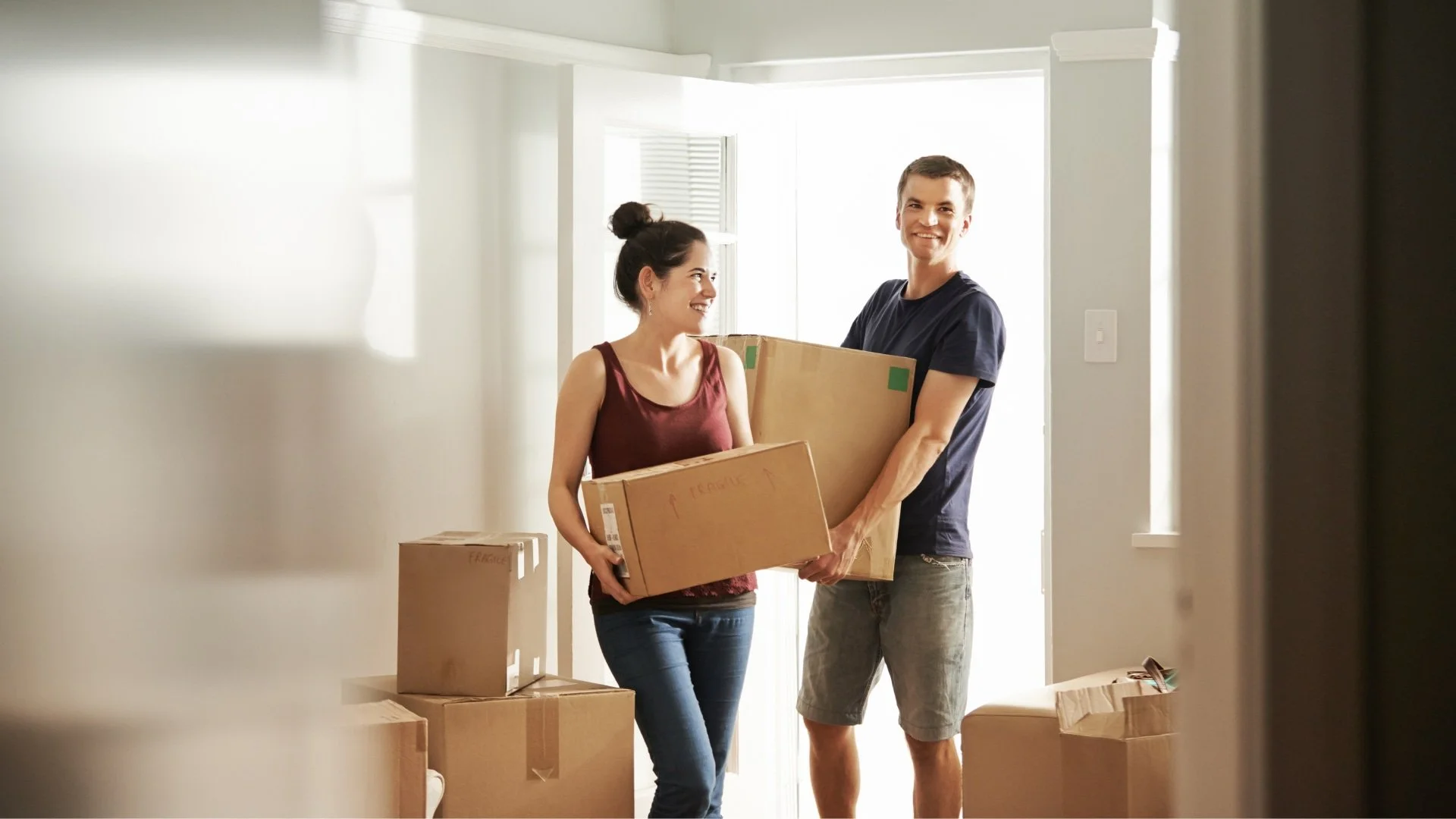 A young couple smiling and carrying moving boxes inside their new home.