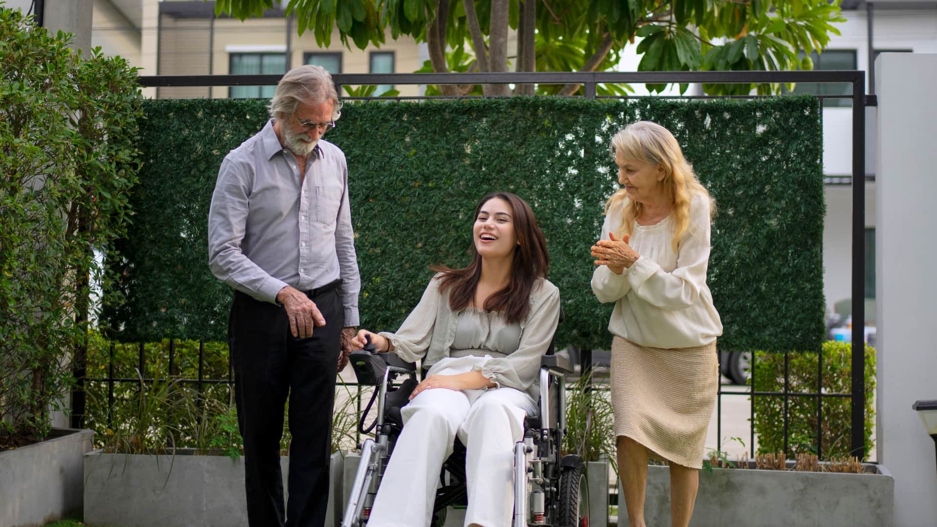 A young woman in a wheelchair outdoors, smiling and laughing, accompanied by an elderly man and an elderly woman, all standing on a patio with greenery and a privacy screen behind them.