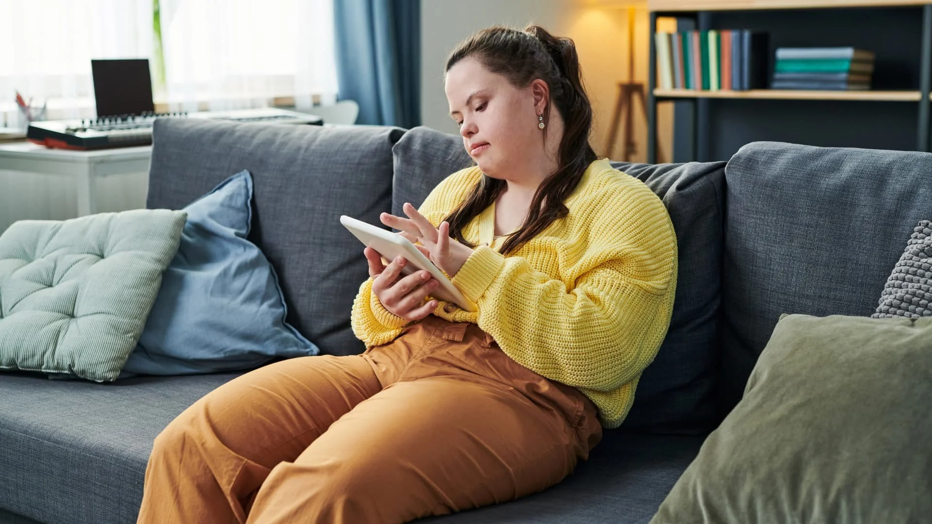 A woman with long dark hair, wearing a yellow sweater and brown pants, sitting on a gray sofa and looking at a white smartphone in her hands in a cozy living room.