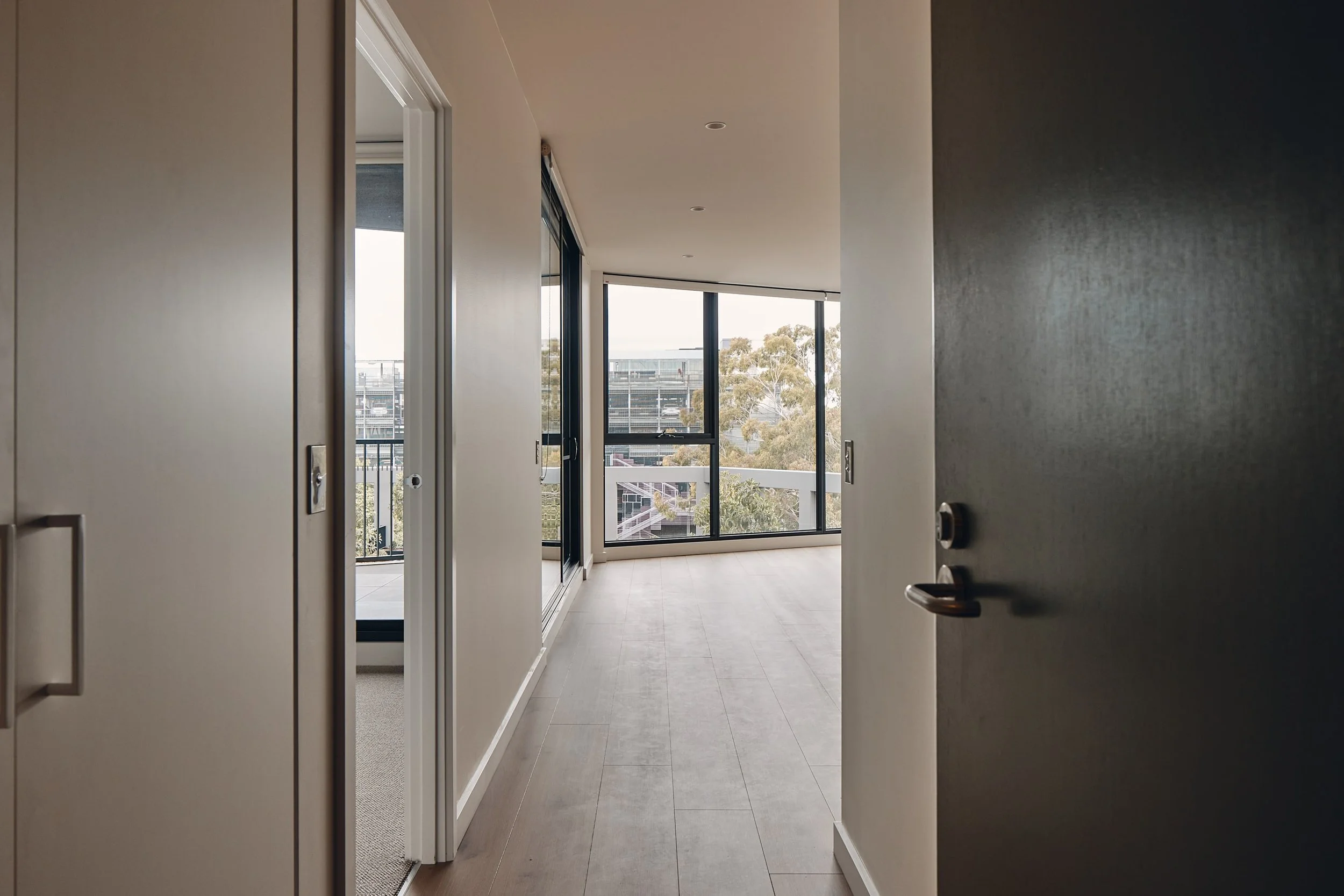 Open door reveals a modern, empty room with large windows, light wood floors, and a view of trees and buildings outside.