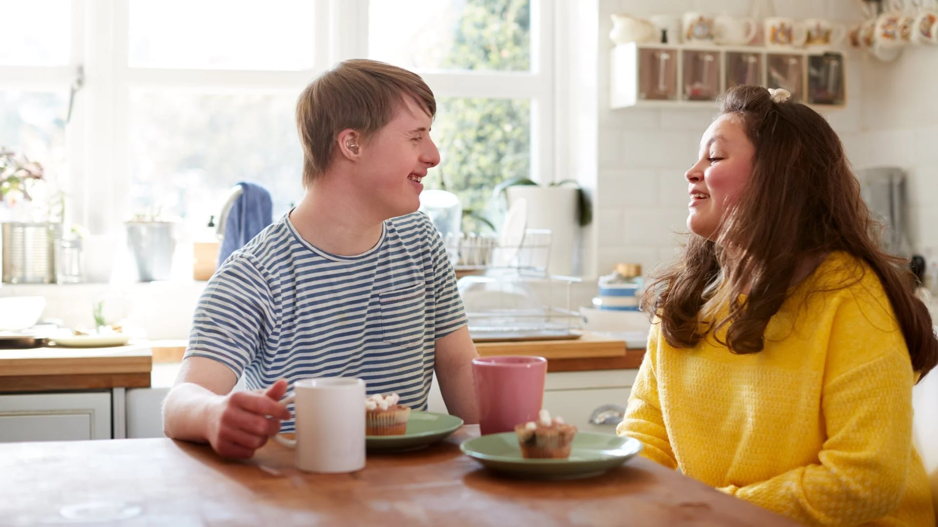 A young man and woman are sitting at a kitchen table, smiling and looking at each other. There are cups and plates with cupcakes on the table. The background shows a bright, sunny kitchen with windows and kitchen shelves.
