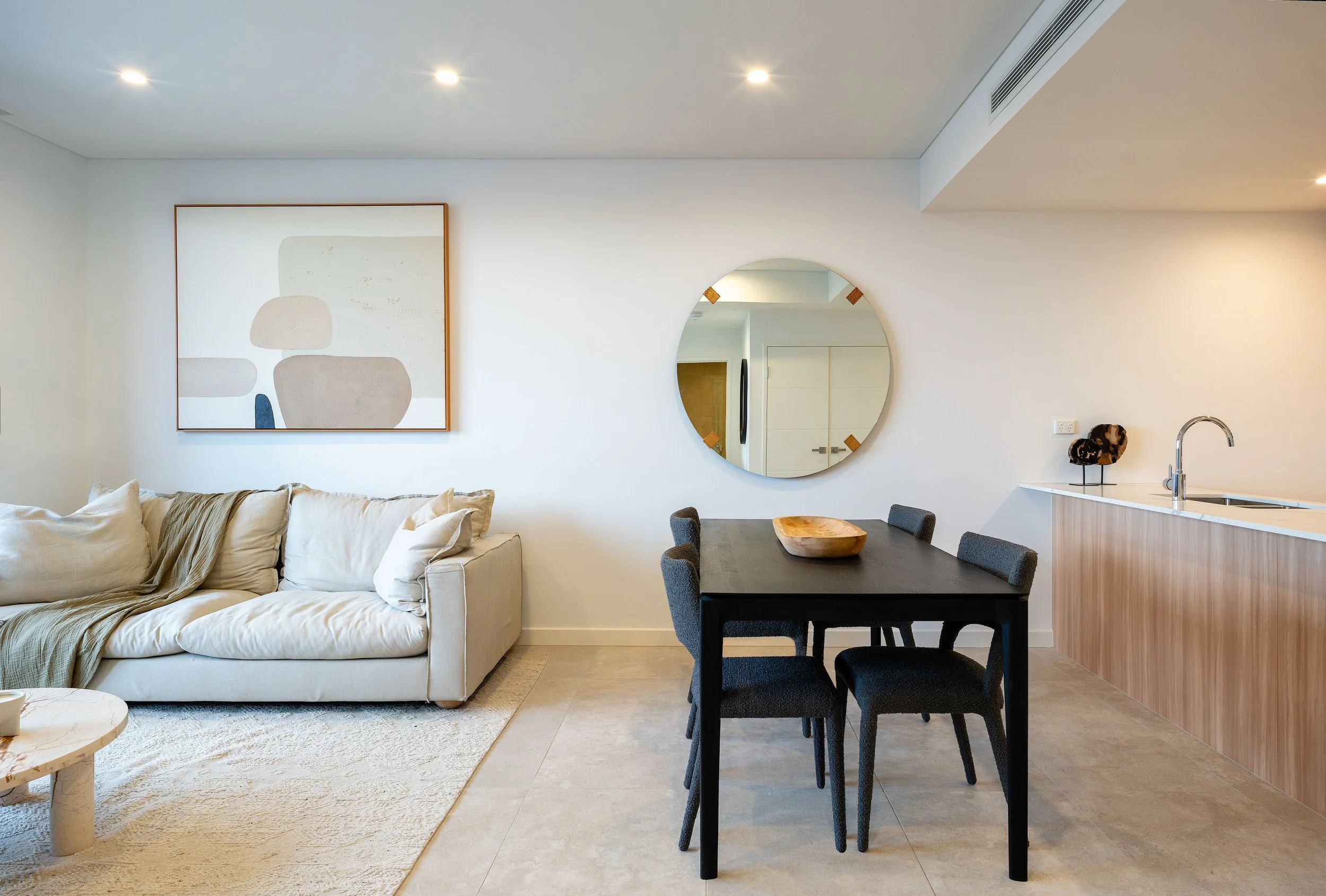 Modern living-dining area with a beige sofa, black dining table, wall art, round mirror, and minimalist decor.