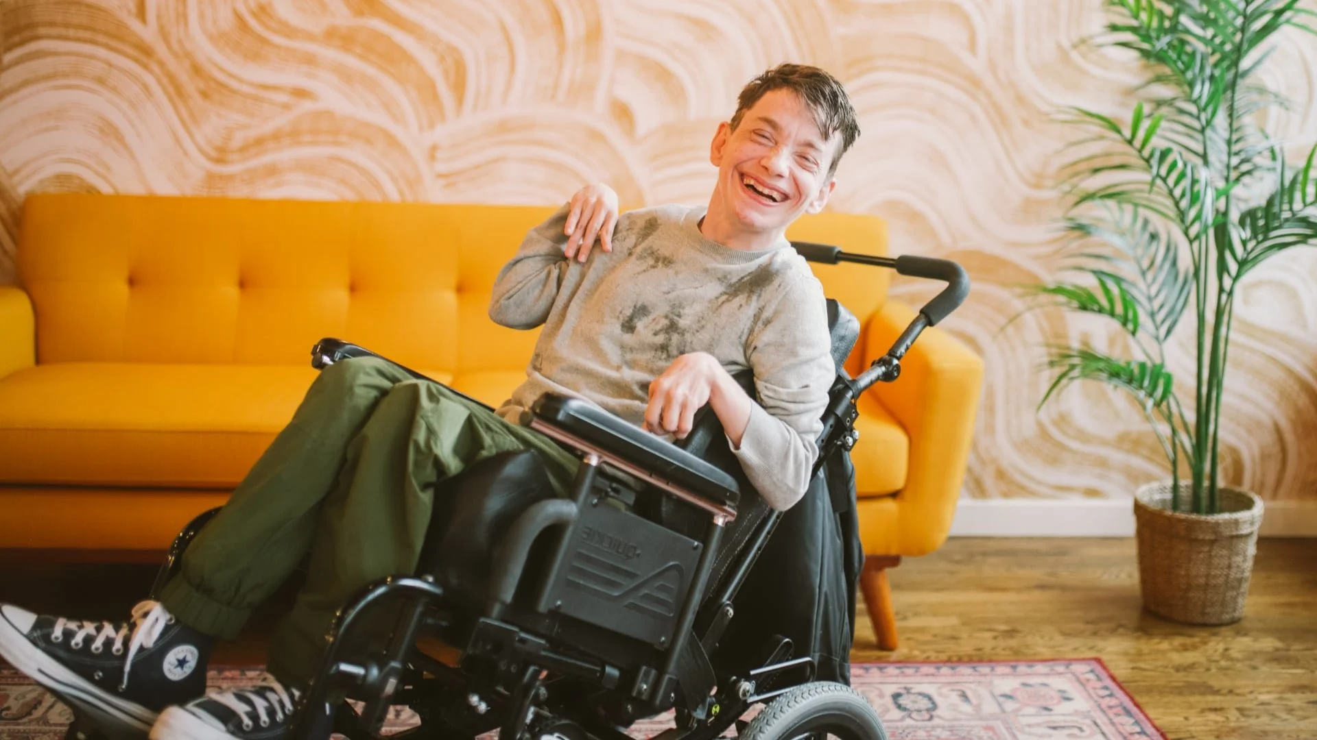 A young man with a big smile sitting in a wheelchair in a cozy living room, with a yellow sofa and a potted plant in the background.
