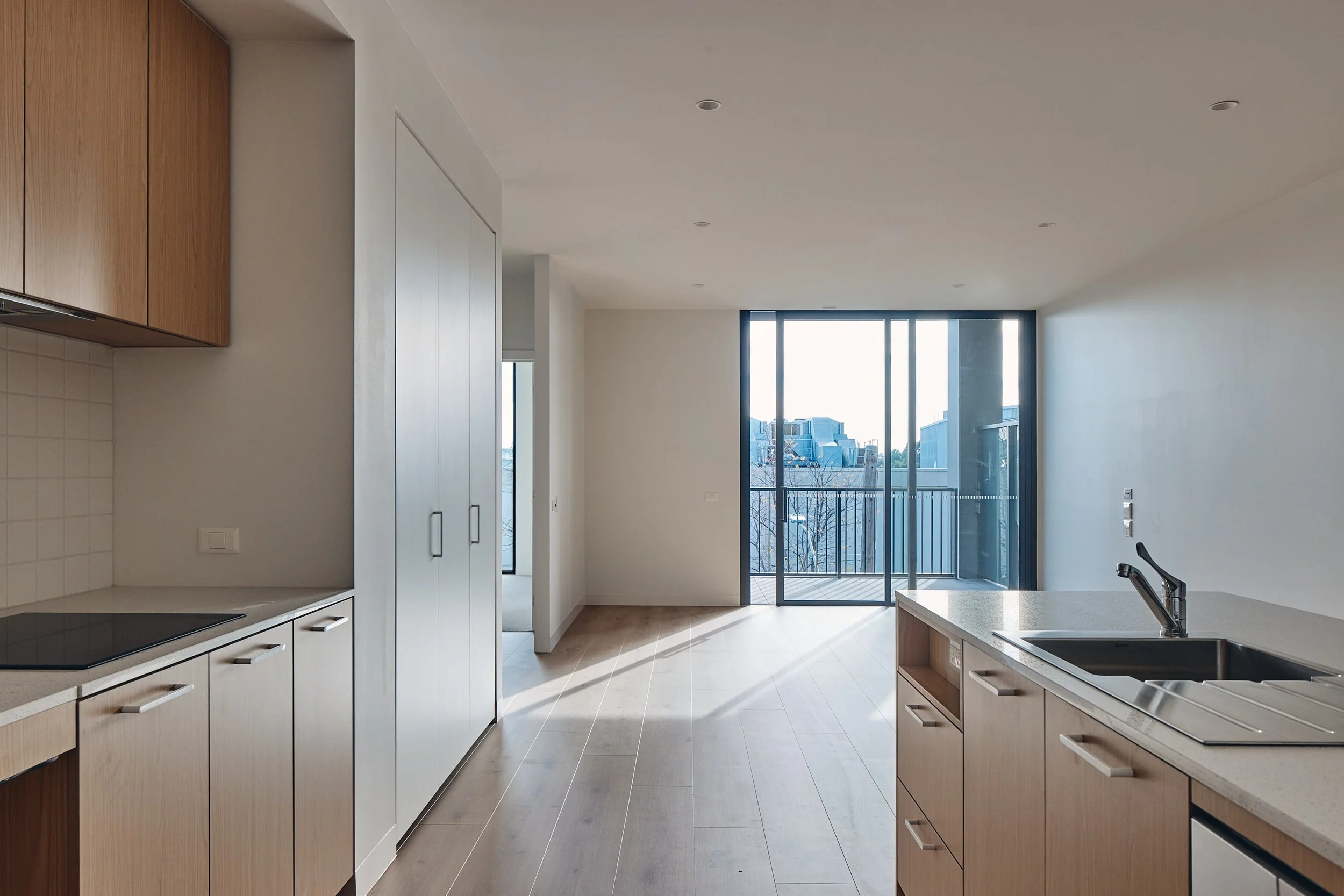 Modern, unfurnished kitchen and living area with light wood cabinets, large windows, and a balcony view.