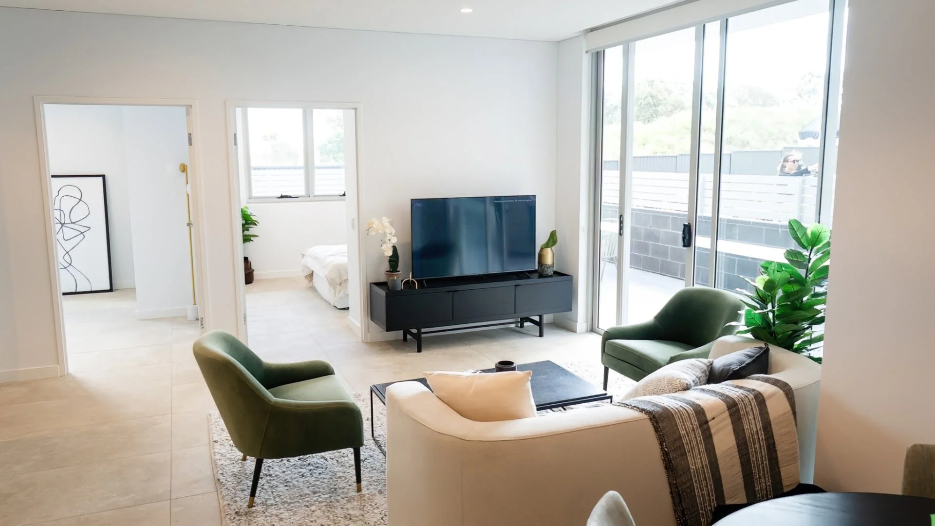 Living room with sliding glass doors, a black TV on a black console, green chairs, a beige sofa with a striped blanket, and indoor plants.