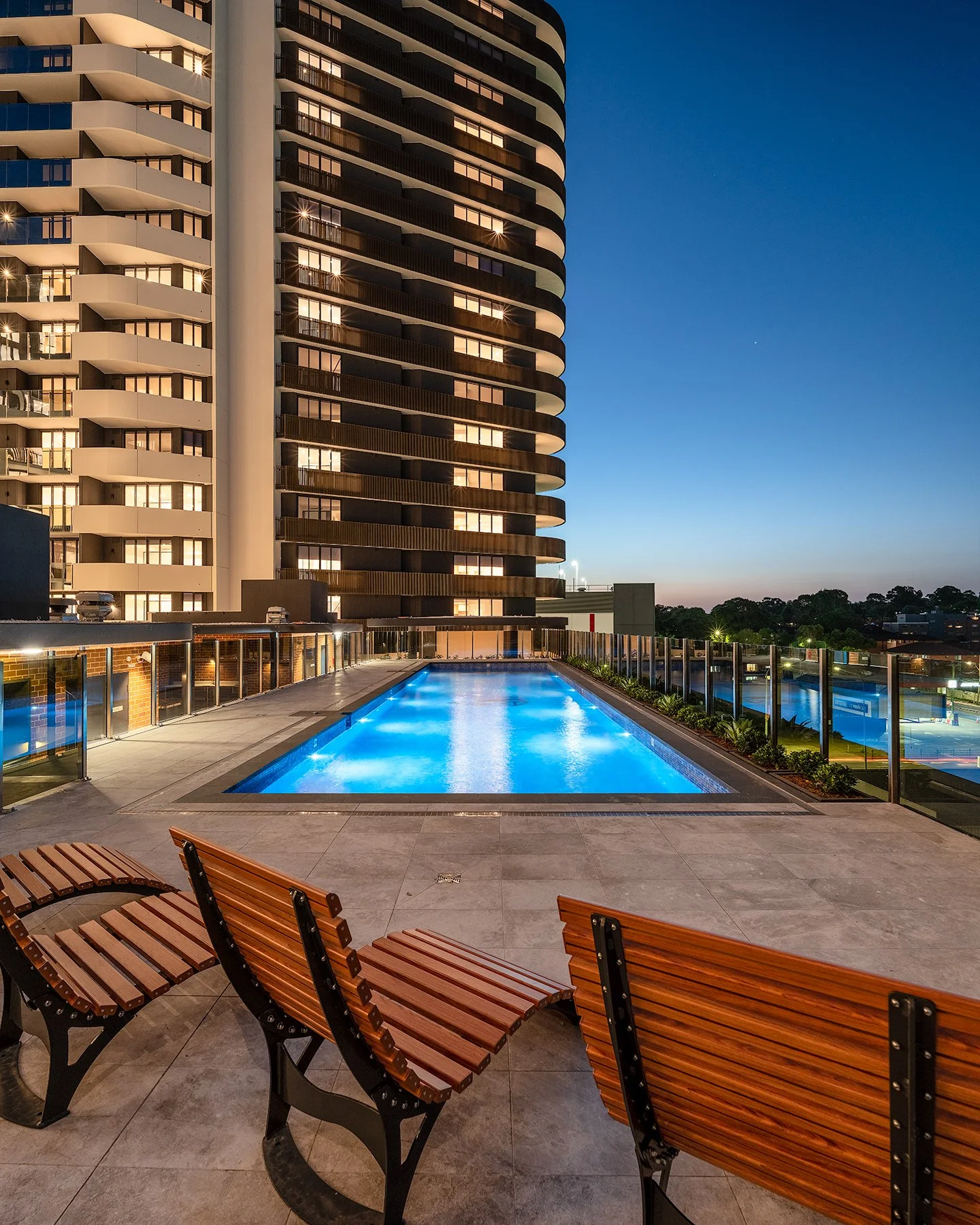 Modern apartment building with illuminated windows and a lit outdoor pool surrounded by lounge chairs at dusk.