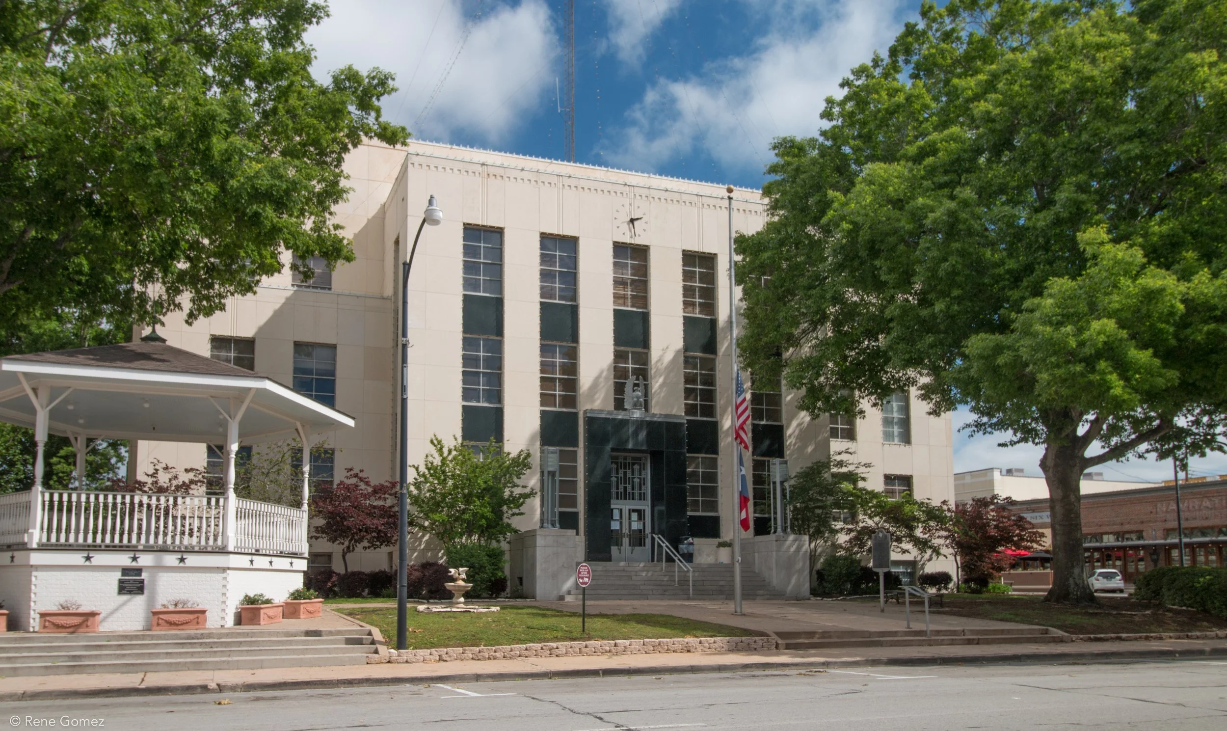 A beige multi-story building with a black entrance, steps leading up to the door, and American and Texas flags in front. Tall green trees and a small gazebo are nearby on a sunny day with a blue sky and some clouds.