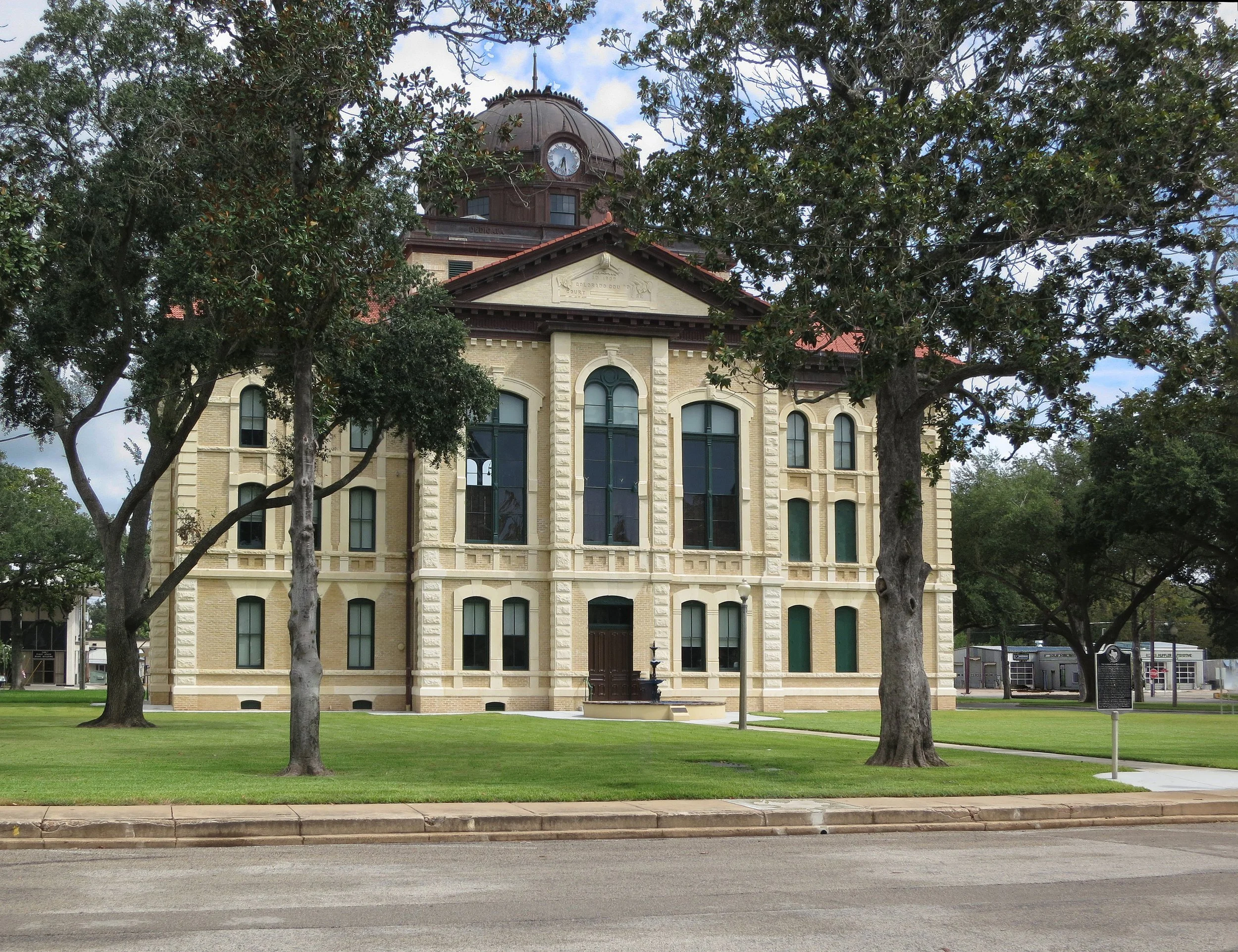 A historic beige brick building with tall arched windows, a clock tower on top, and surrounded by green trees and grass.