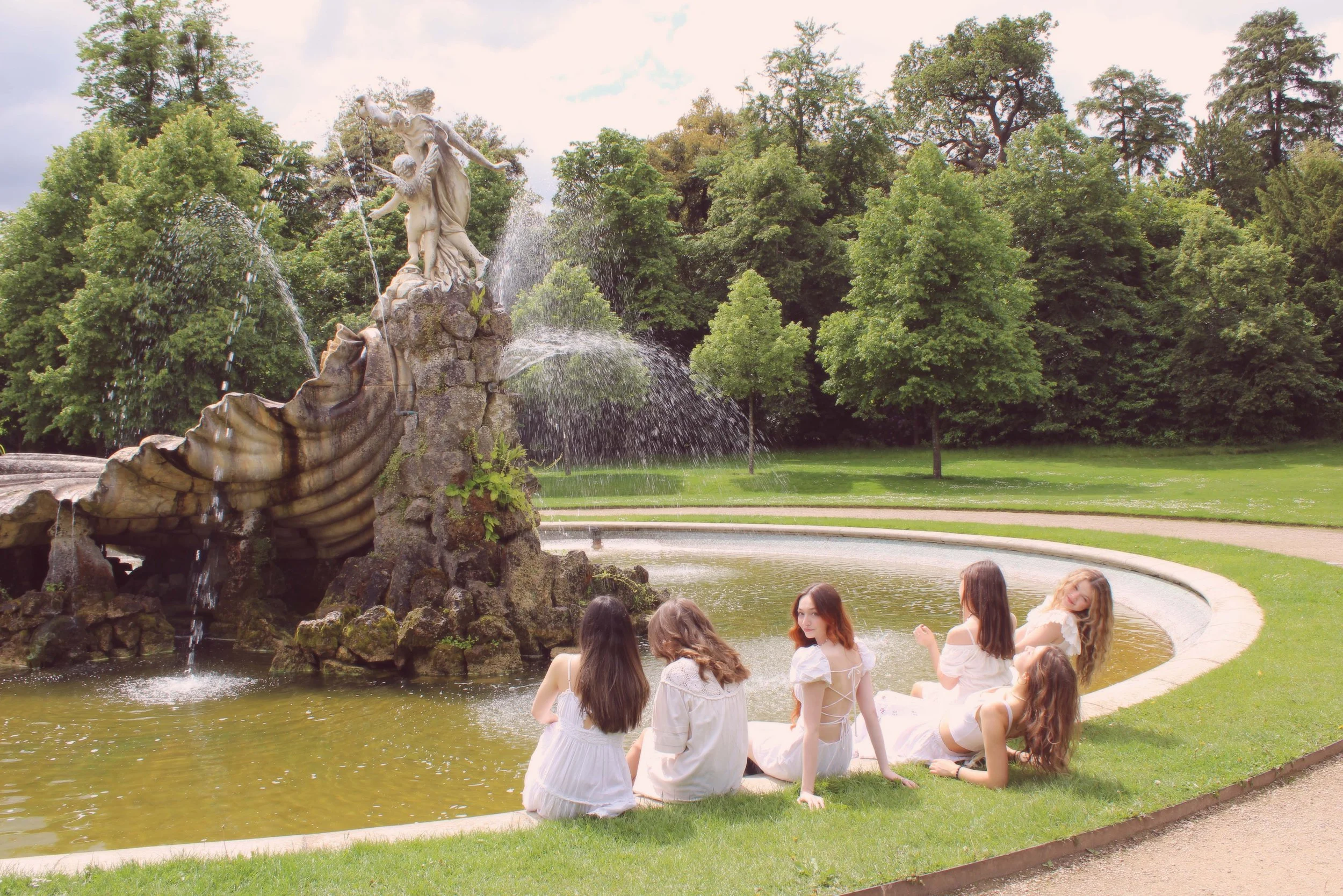 Six young women in white dresses sitting by a circular fountain with a large ornate sculpture of two figures, water spraying from the sculpture, surrounded by green trees and grass.