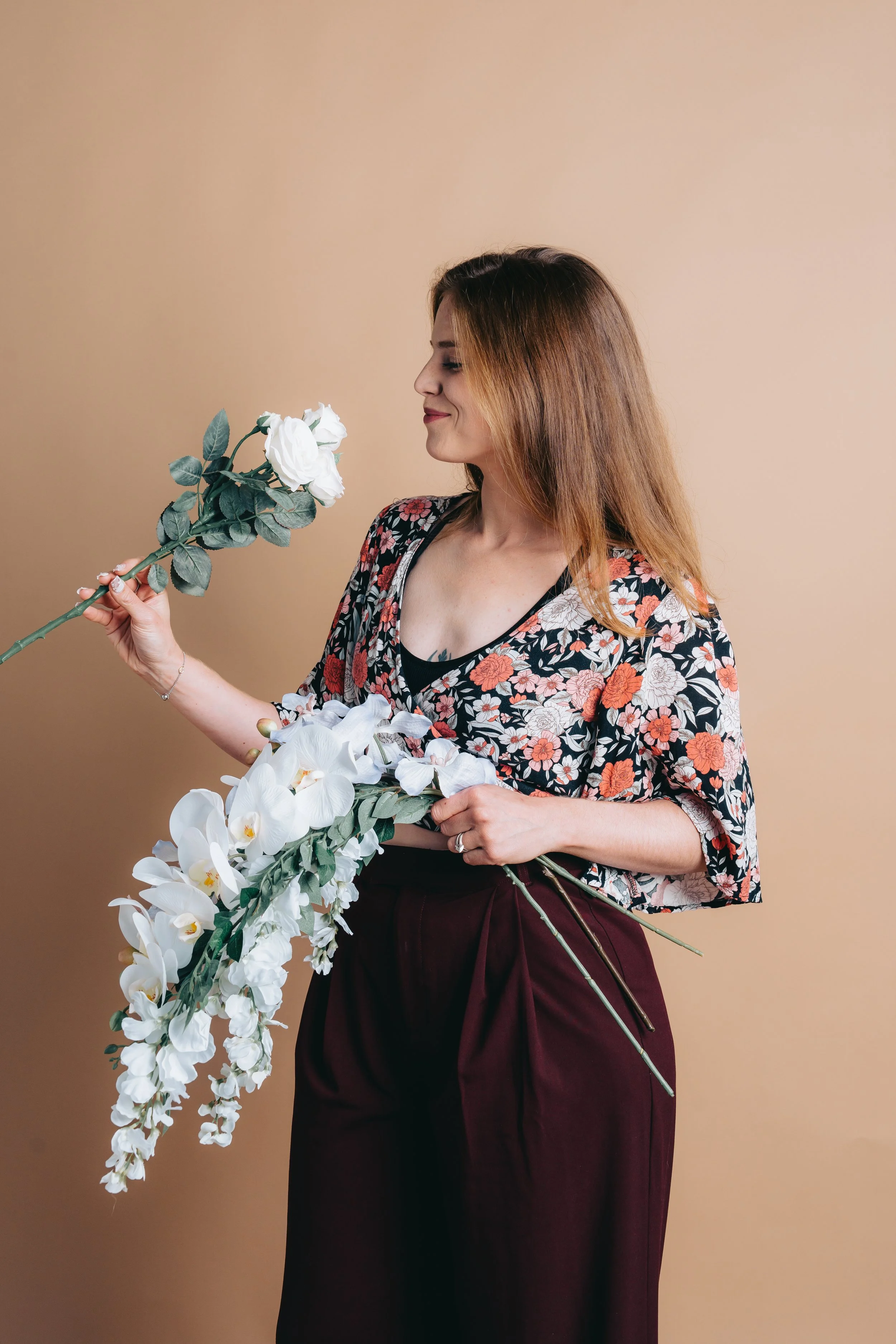 A woman with long brown hair wearing a floral blouse and maroon pants holding a bouquet of white orchids and gazing at a single white rose.