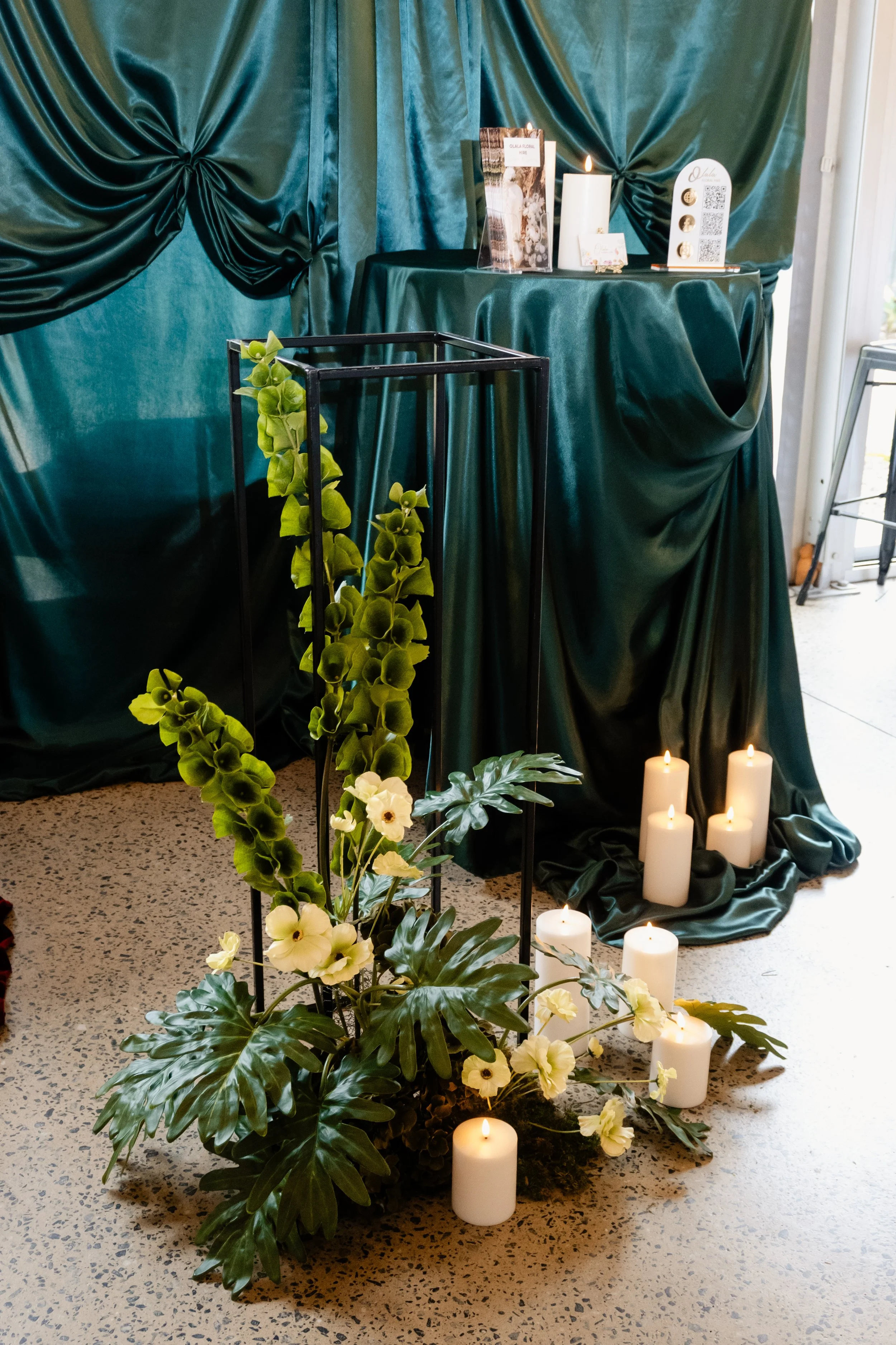 A dark green satin-covered table with white candles, a decorative display with a photo, and QR codes. In front, a white and green floral arrangement with candles on the floor.