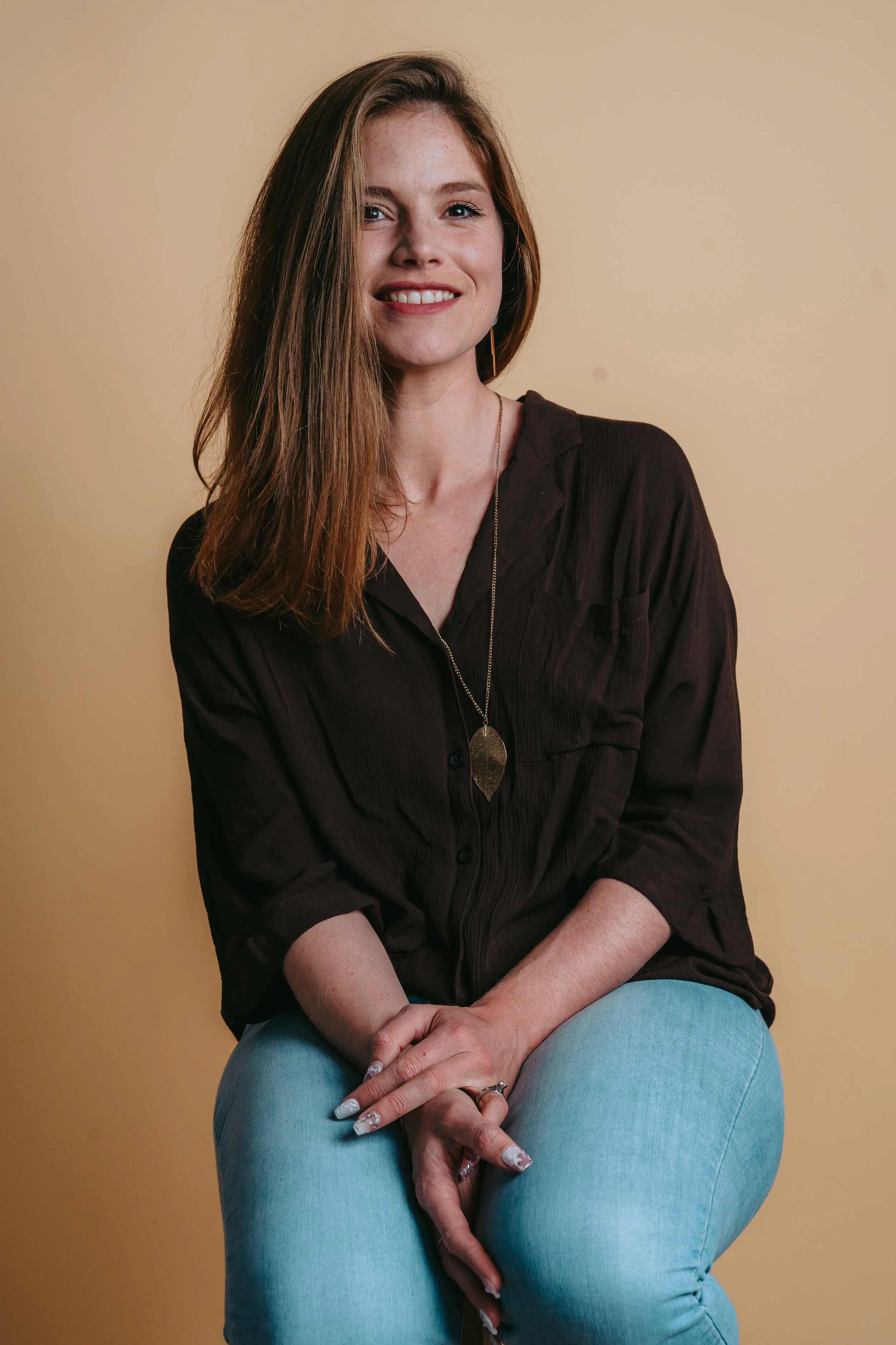 A woman with long reddish-brown hair, wearing a dark brown shirt and light blue jeans, sitting against a plain yellow background and smiling at the camera.
