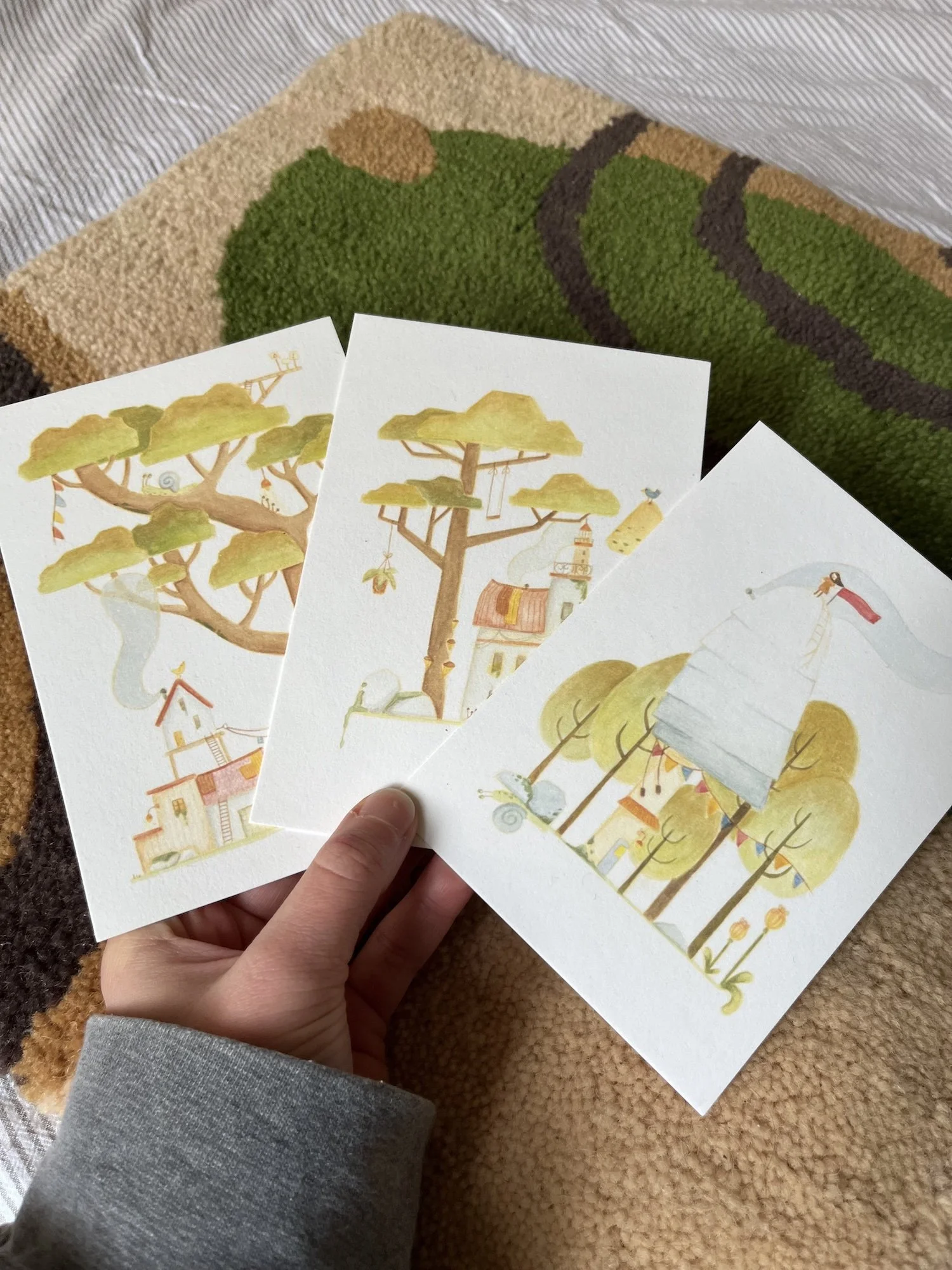 Three illustrated greeting cards featuring whimsical trees and houses, held by a person's hand over a textured rug with a tree design.