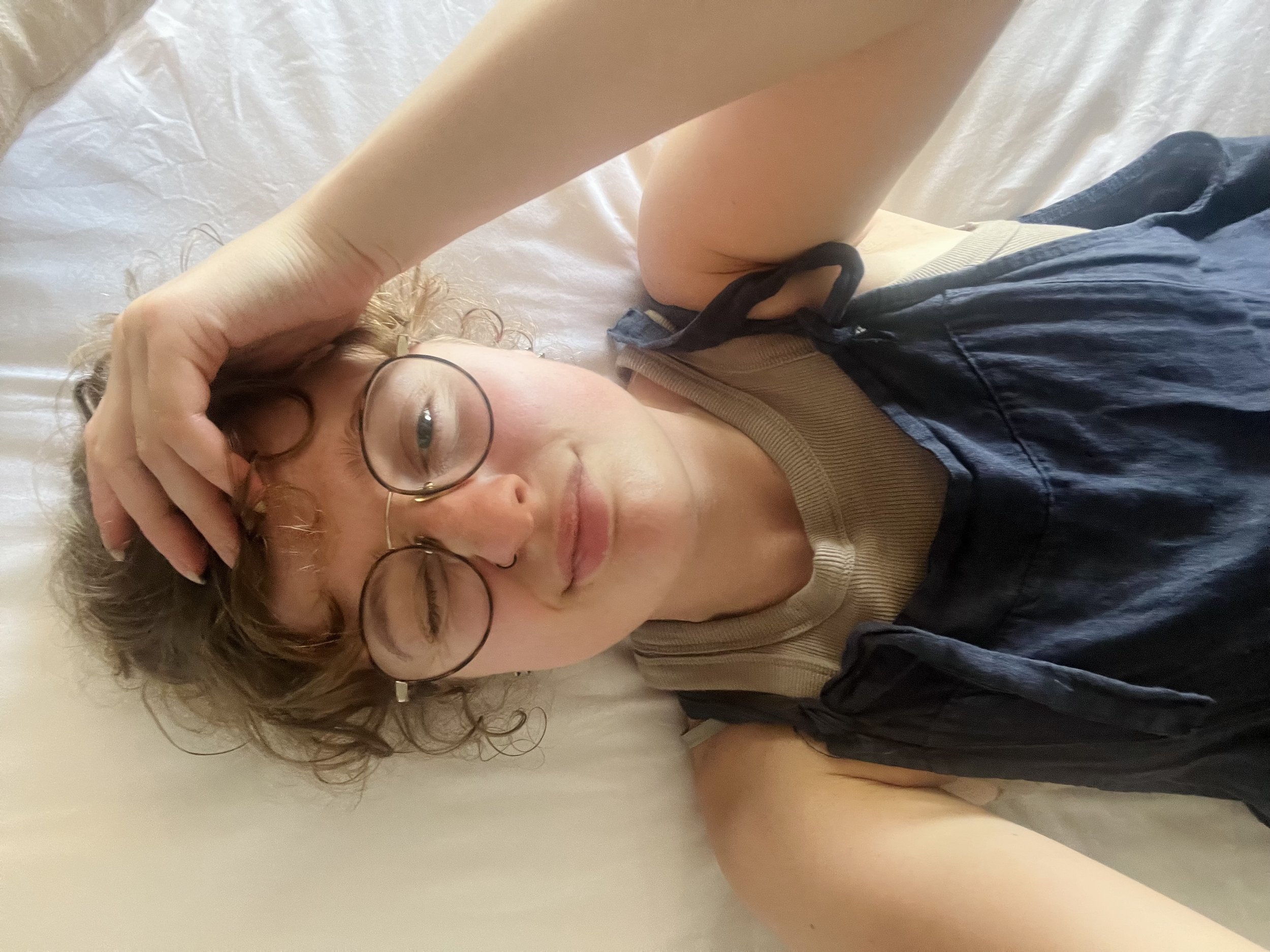 A woman with glasses lying on her back on a bed, with her hand on her forehead and her eyes closed.