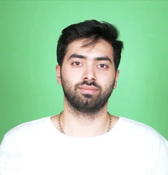 Portrait of a young man with dark hair and beard wearing a white shirt and gold chain against a green background.