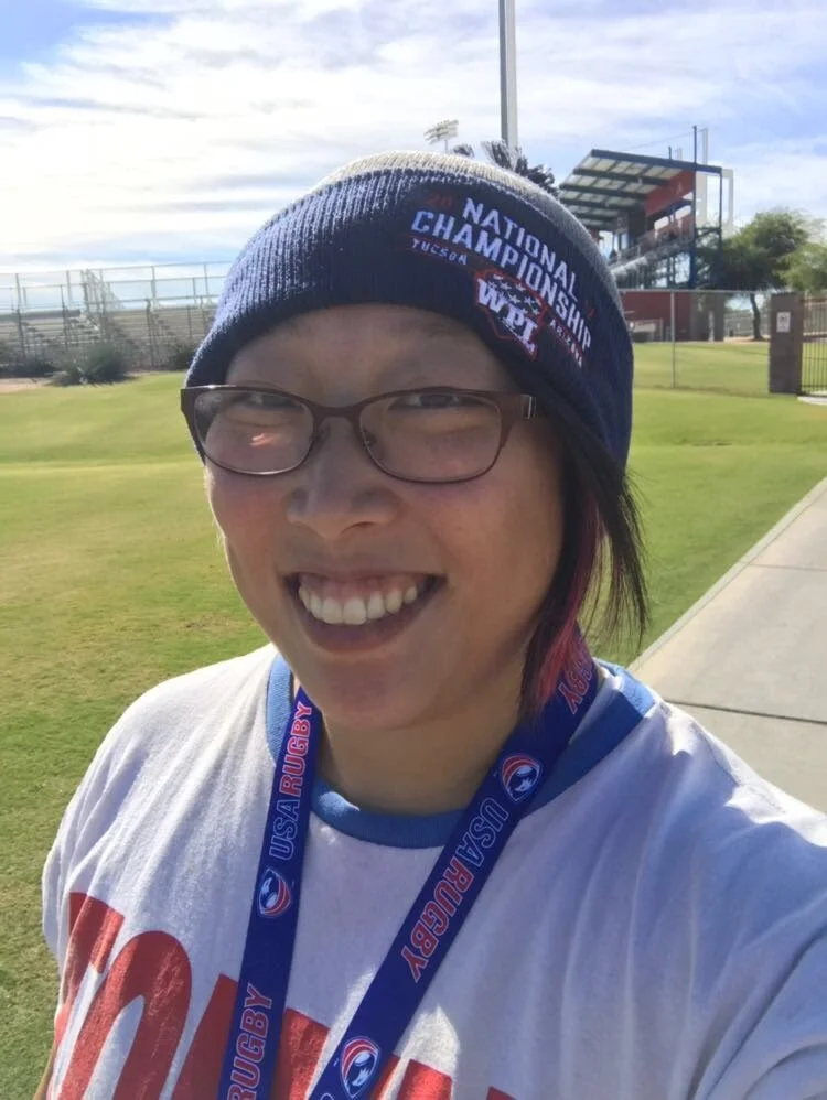A young asian woman wearing a beanie and a lanyard, smiling, with grass and blue sky in the background.
