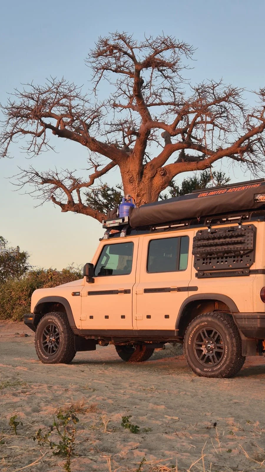 A beige off-road vehicle parked on a dirt road, with a large leafless tree in the background during sunset.