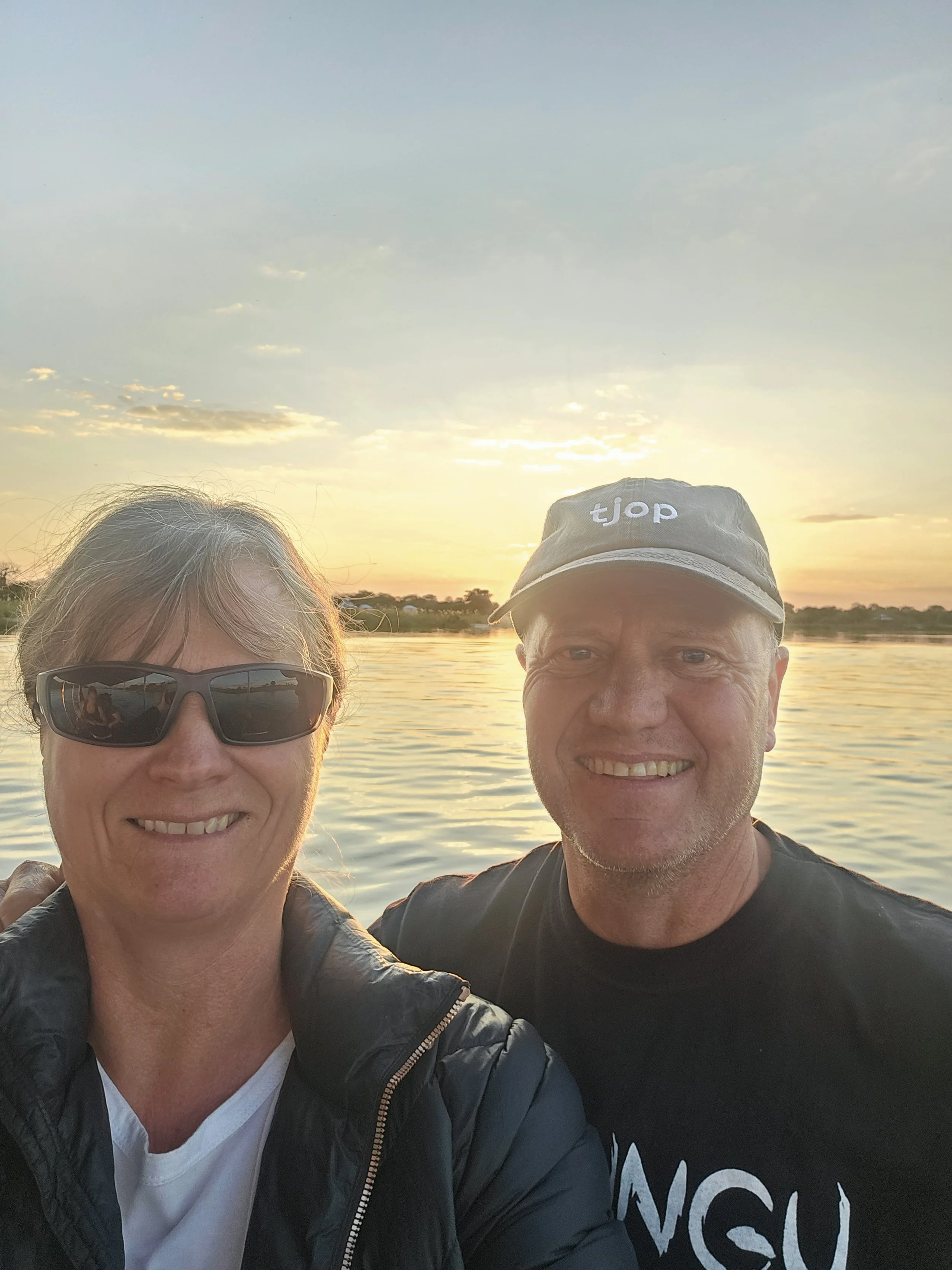 A smiling man and woman taking a selfie by a body of water at sunset. The woman is wearing sunglasses and a black jacket, and the man is wearing a black t-shirt and a cap that says 'tjop'.