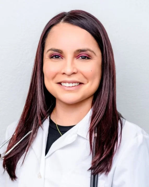 Healthcare professional with long dark hair wearing a white coat and stethoscope, smiling against a plain light gray background.