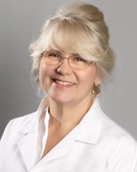 A smiling woman with blonde hair styled in a bun, wearing glasses, gold hoop earrings, and a white collared shirt, against a gray background.
